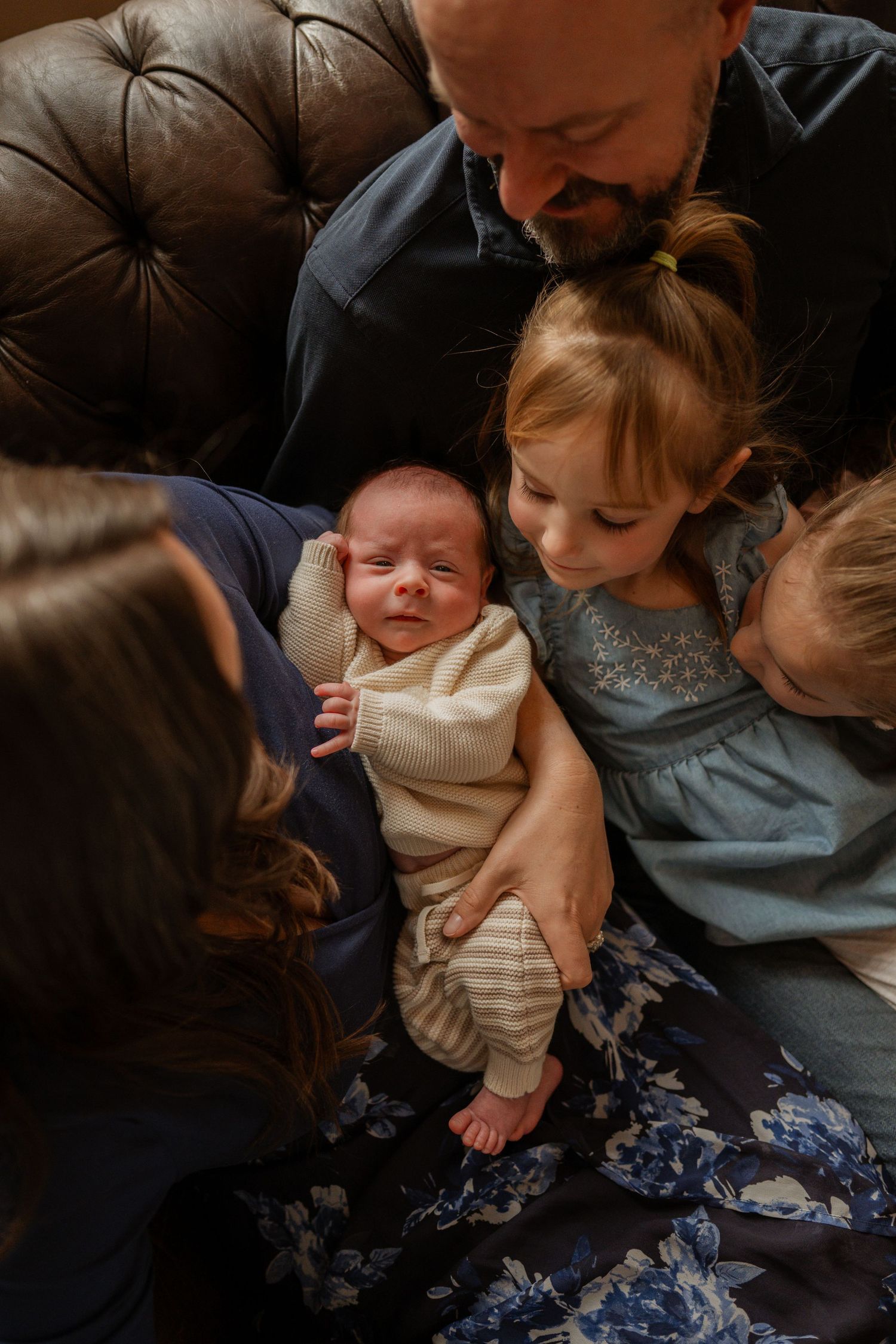 A family sits together on a dark leather couch while holding and interacting with a small baby.