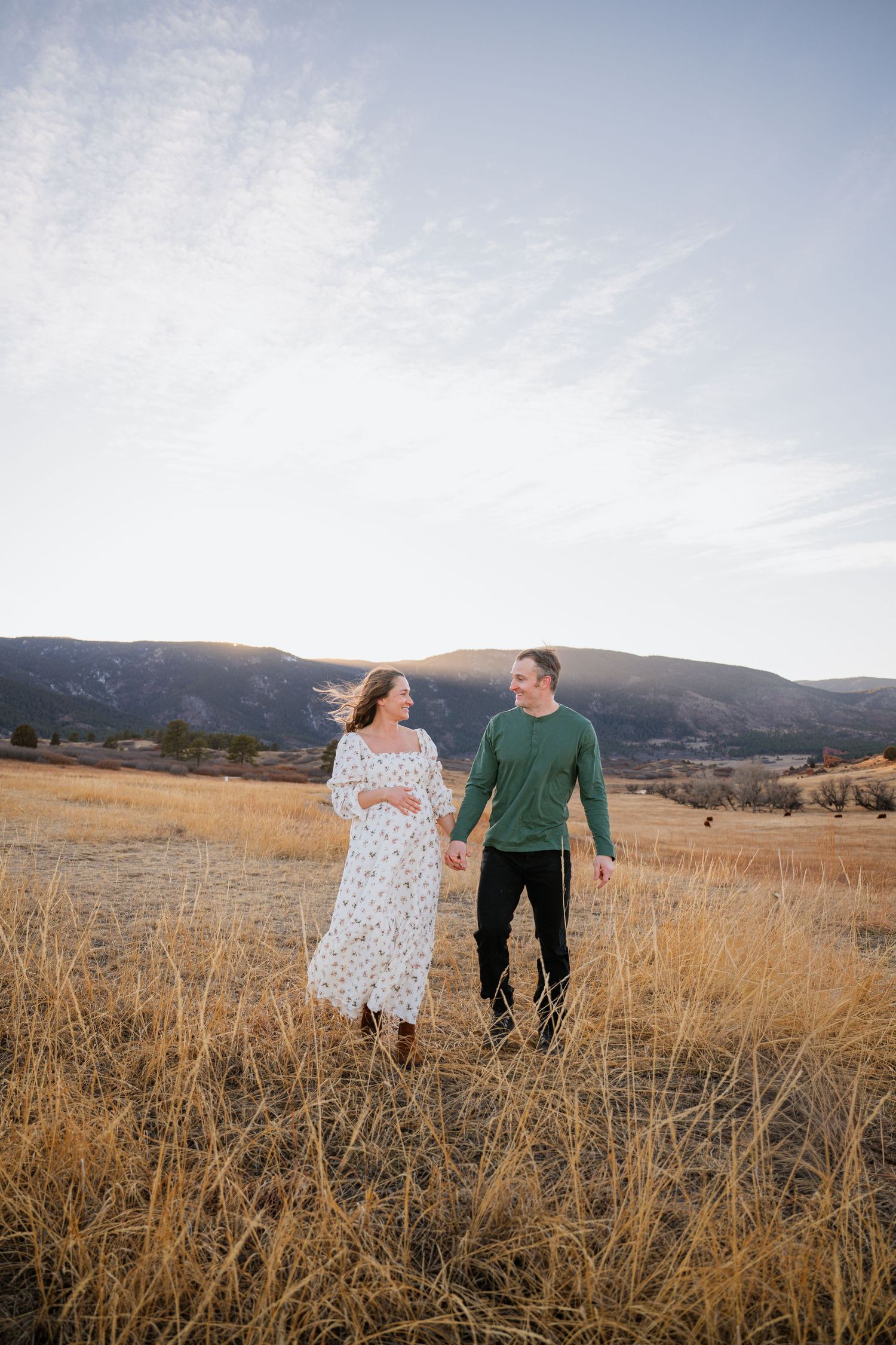 A couple walks together through a golden autumn field with mountains in the background during sunset.