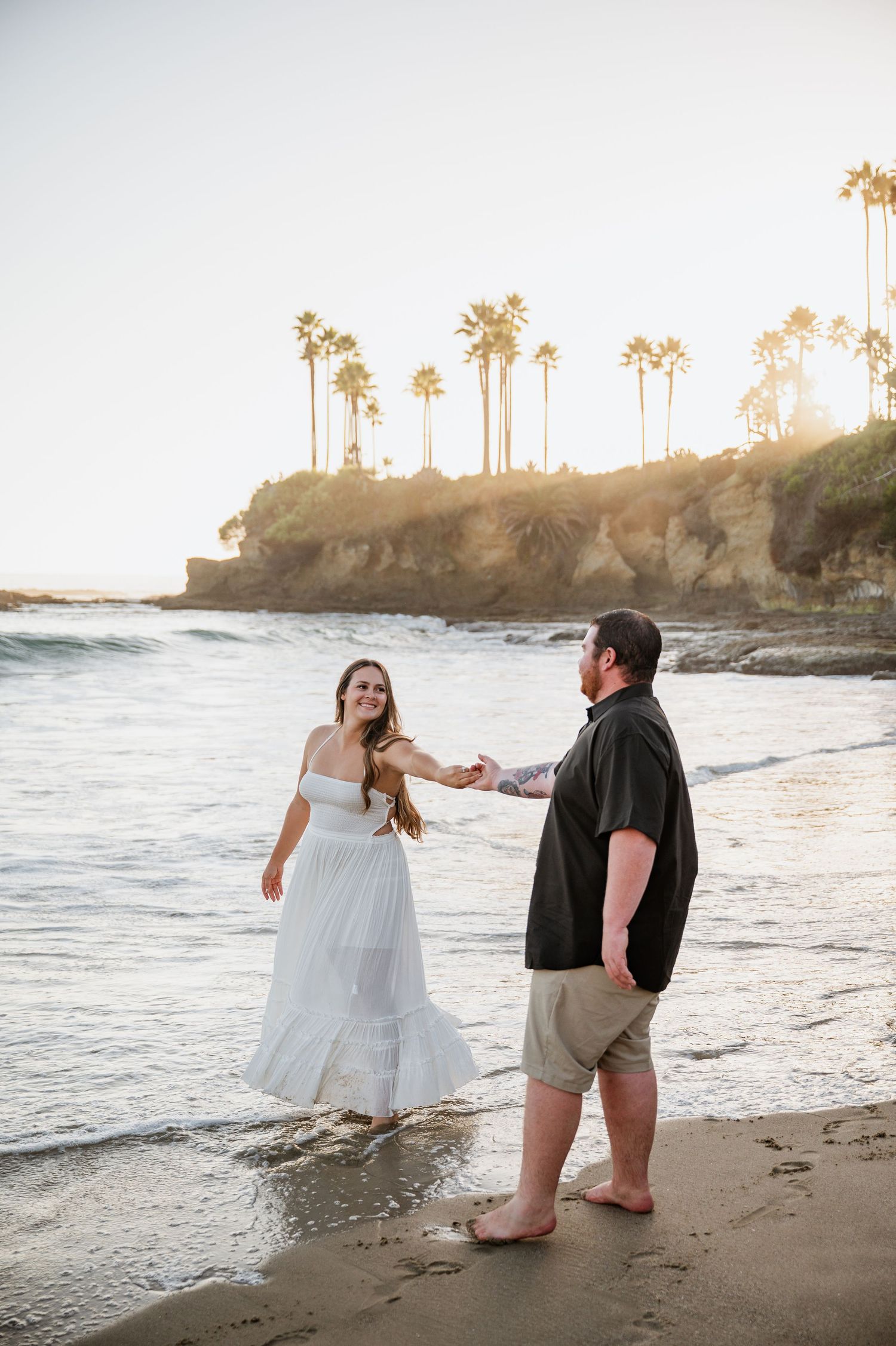 A couple in wedding attire walks along a sunset beach with palm trees in the background.