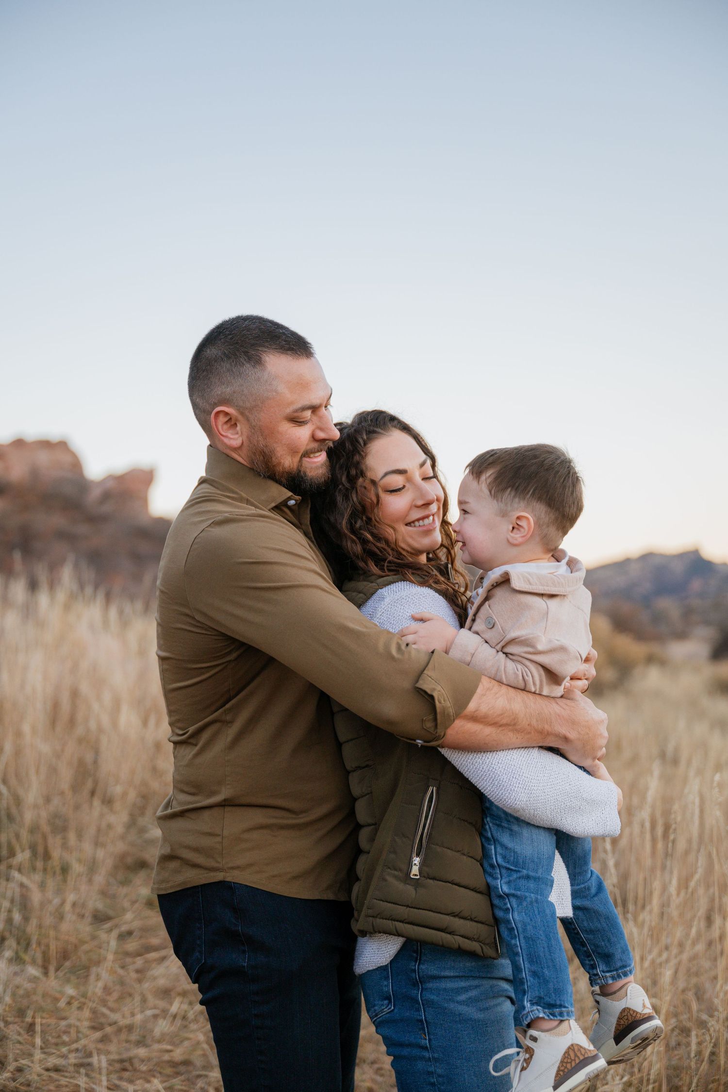 A family shares a tender embrace while standing in a field of tall grass during golden hour with mountains in background.