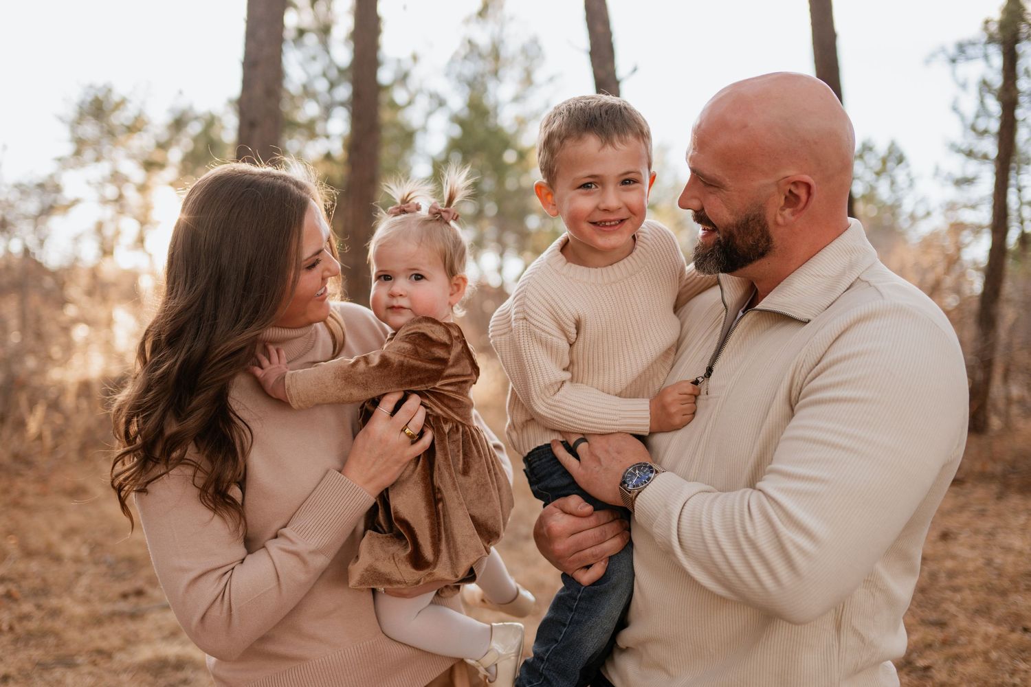 A family wearing neutral colors shares joyful moments together during an outdoor photo session in a wooded setting.