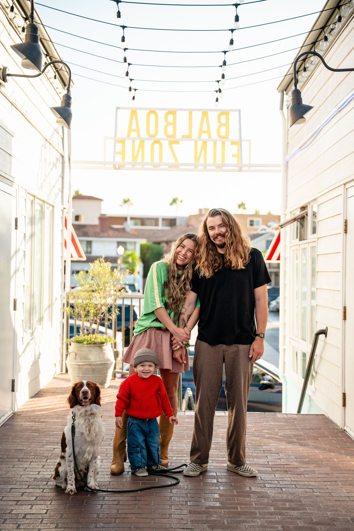A family and their dog stand together under string lights on a brick walkway outside a storefront.
