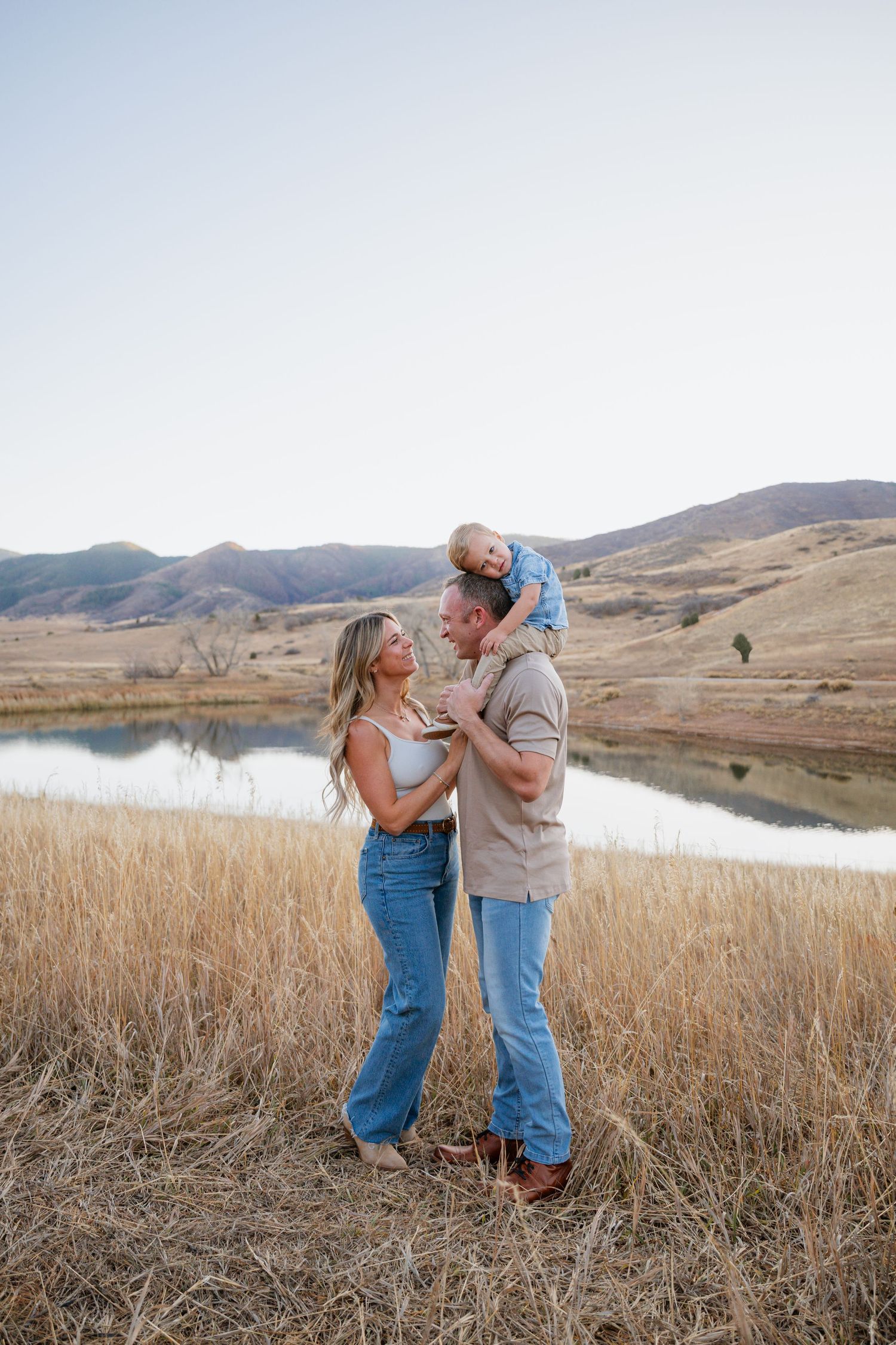 Couple shares romantic moments in golden field by mountain lake during sunset photo session.