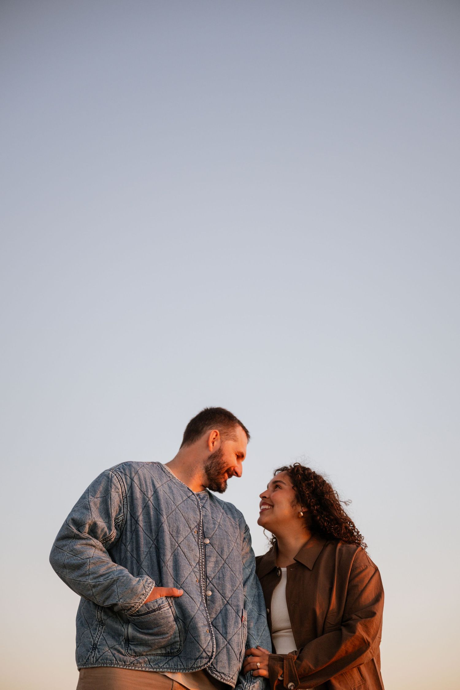 A happy couple laughing together outdoors during sunset while wearing casual denim and brown outfits.
