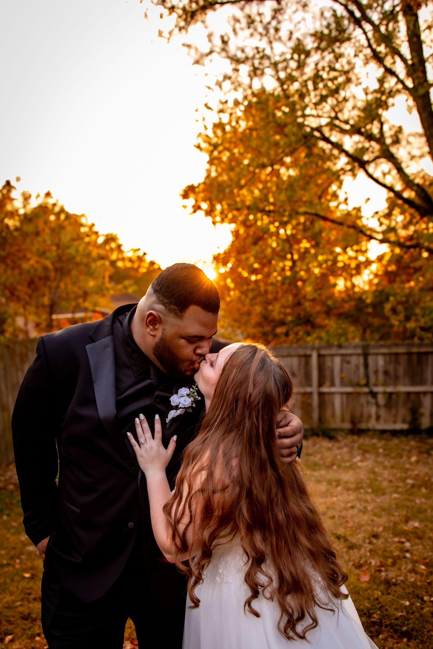 Couple shares romantic kiss in autumn backyard with golden sunset light filtering through trees and fallen leaves.