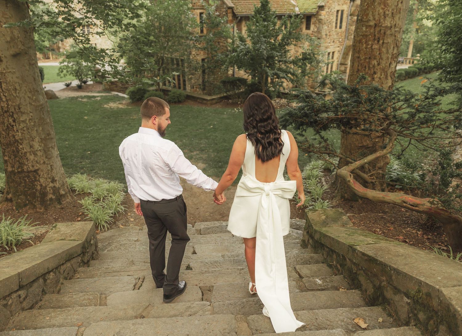 Two people walk hand in hand down a stone garden path surrounded by trees and greenery.
