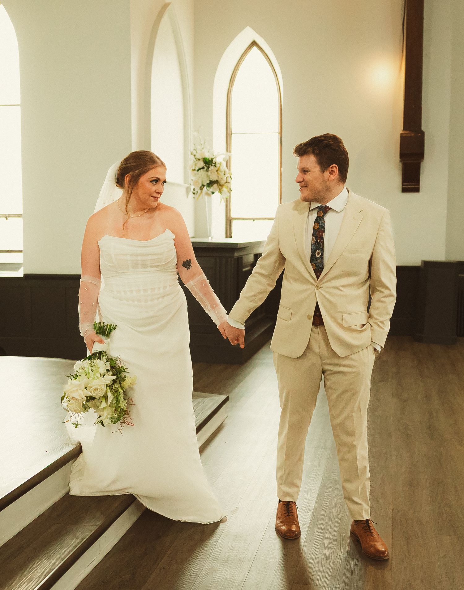 A newlywed couple holds hands in a bright church interior with gothic arched windows and wooden floors.