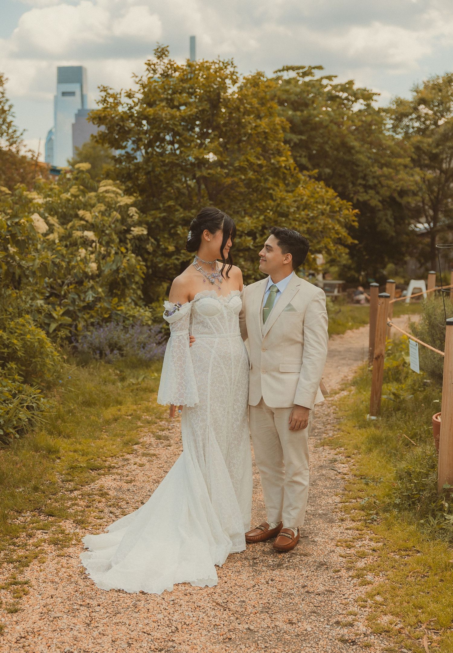 A couple in wedding attire poses together on a gravel path surrounded by green trees and foliage.