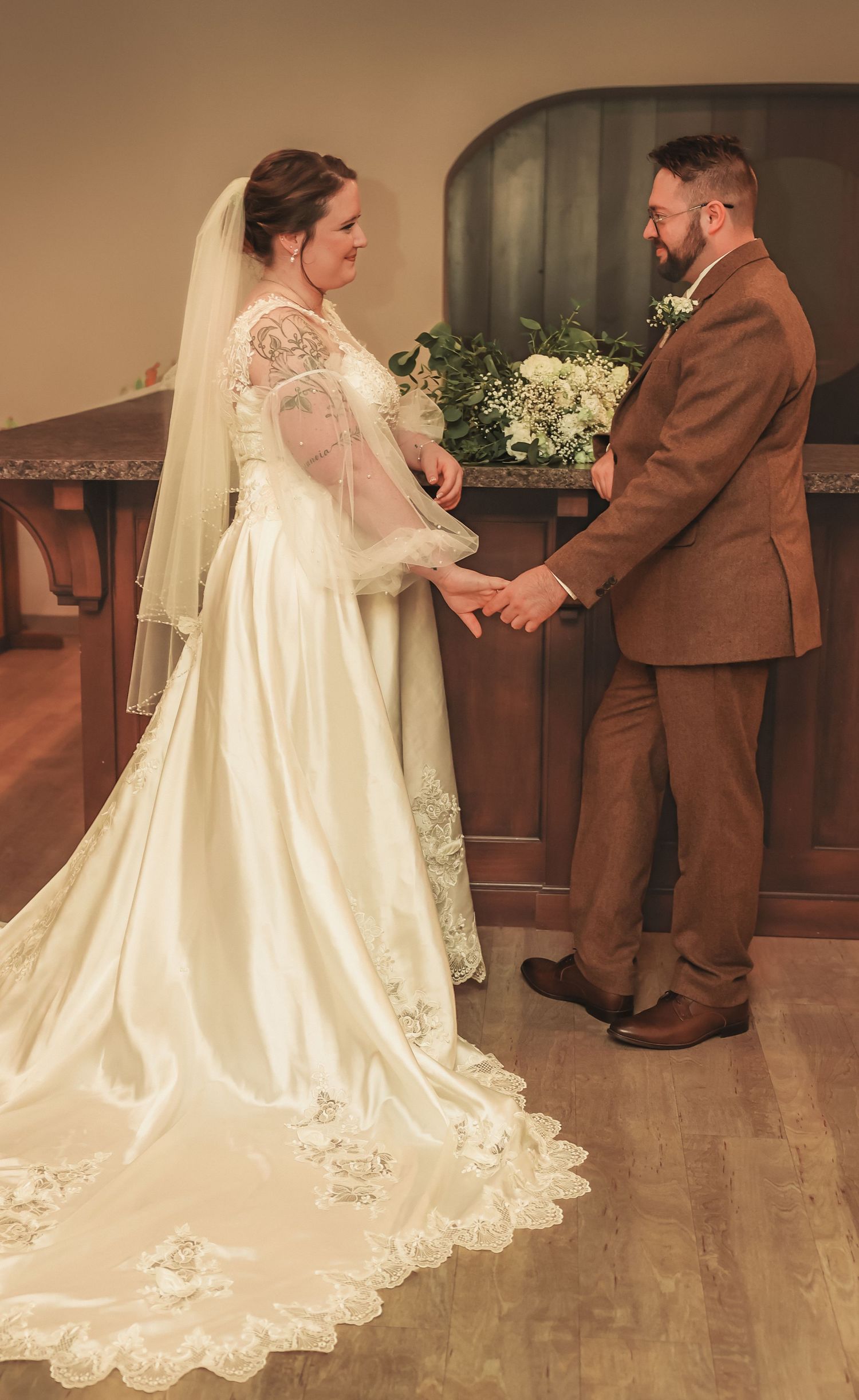 A couple exchanges wedding vows in a chapel, with the bride wearing a lace-trimmed white gown and the groom in a brown suit.