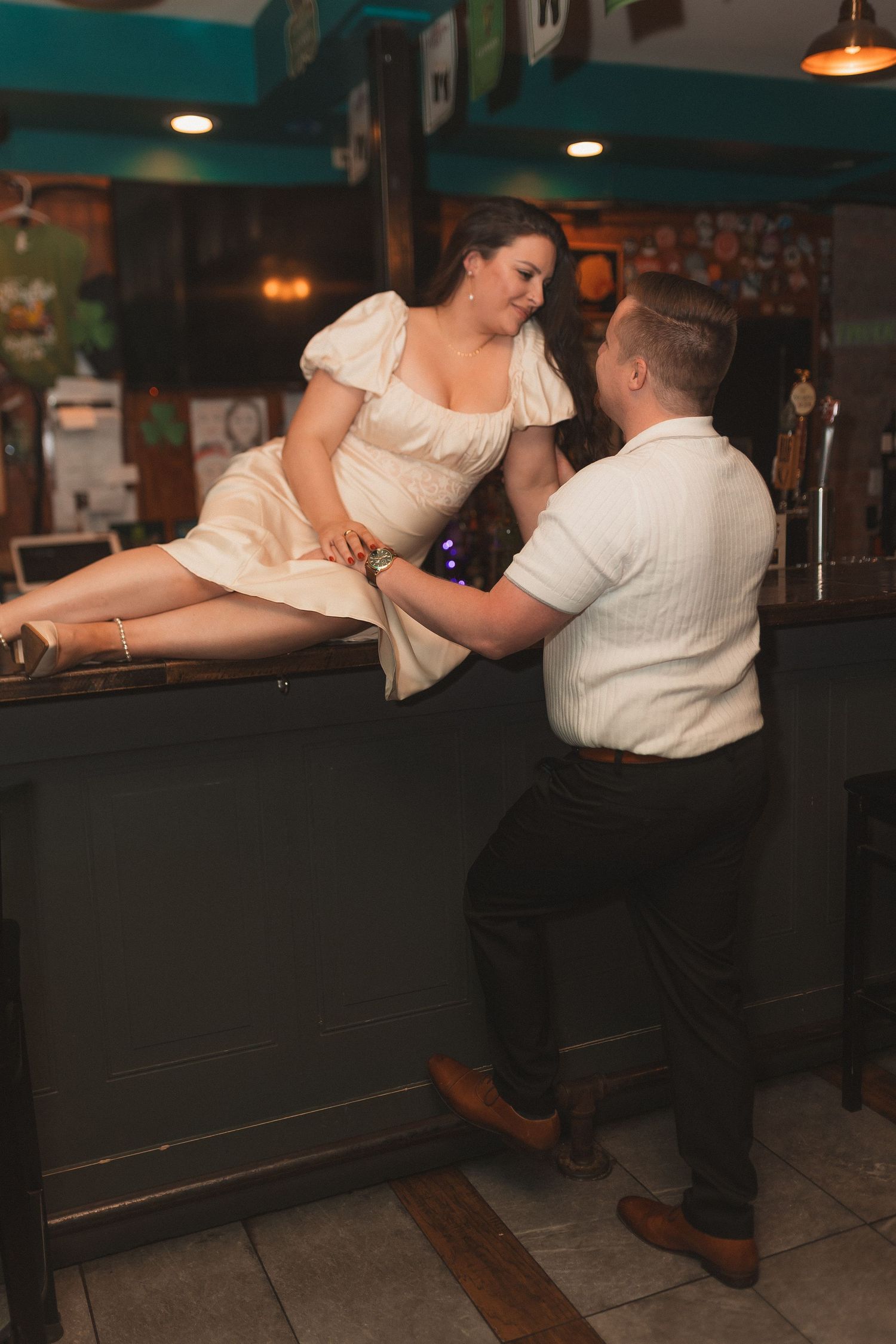 A couple in vintage-style clothing share a romantic moment at a retro-themed bar counter in soft lighting.