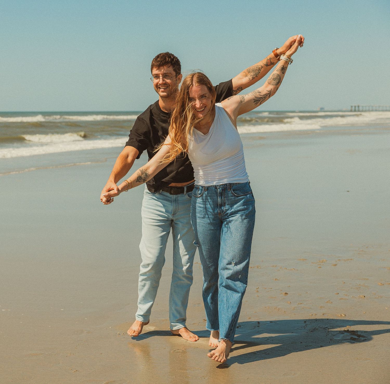 Two people wearing jeans stroll barefoot along a sunny beach shore while playfully posing together.