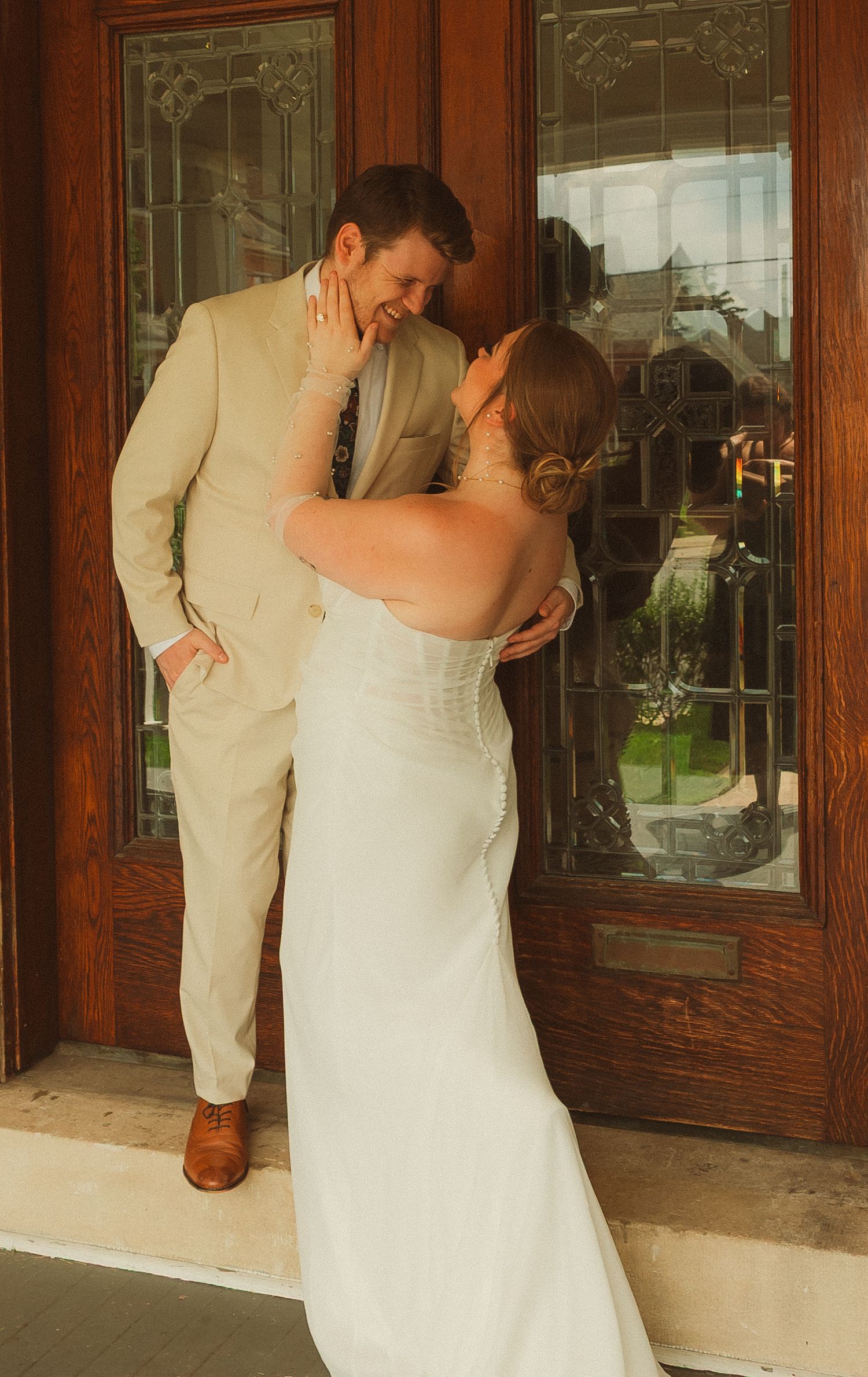 A couple in wedding attire shares an intimate kiss on the porch of a building with wood paneling and glass windows.