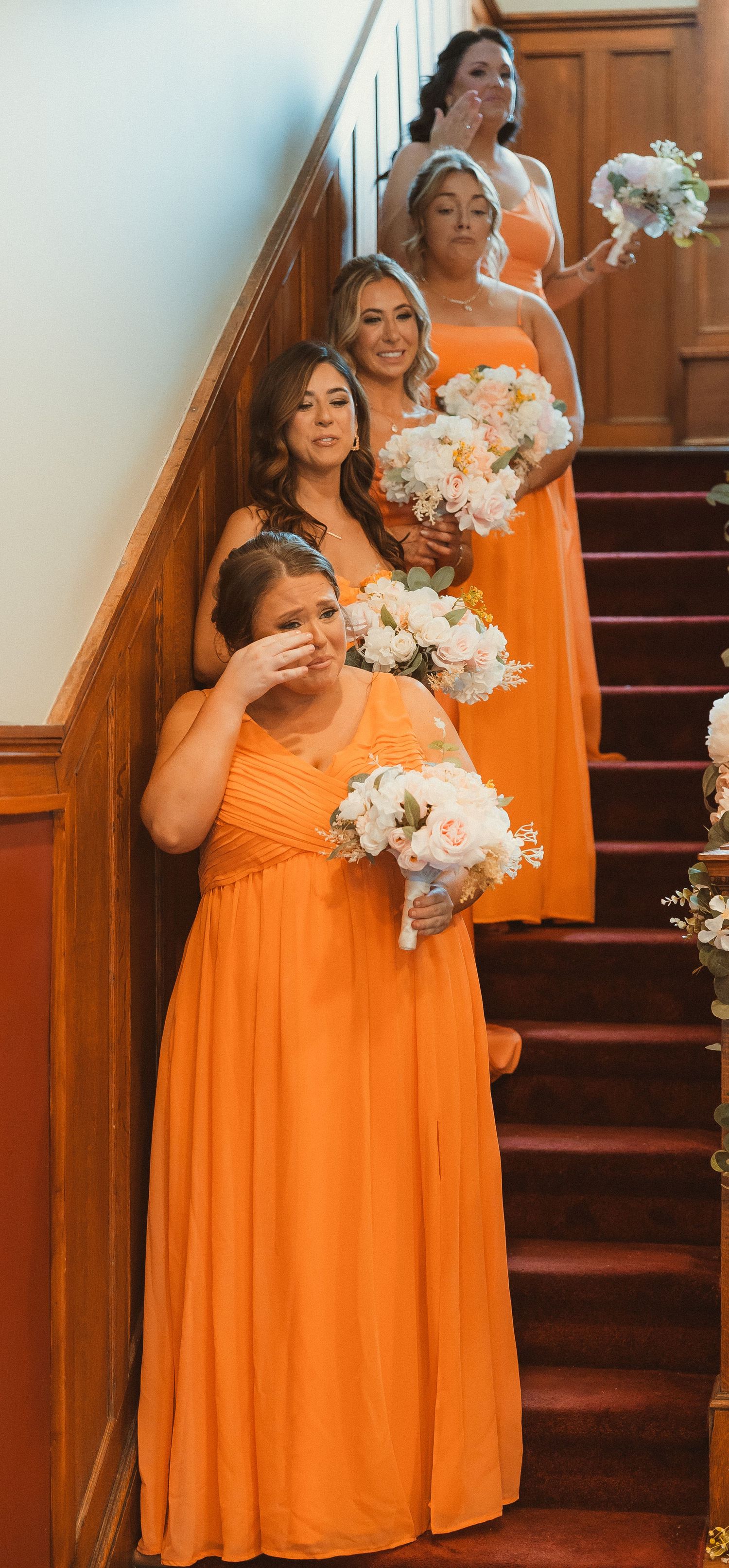 Bridesmaids in bright orange dresses holding white flower bouquets pose on wooden staircase.