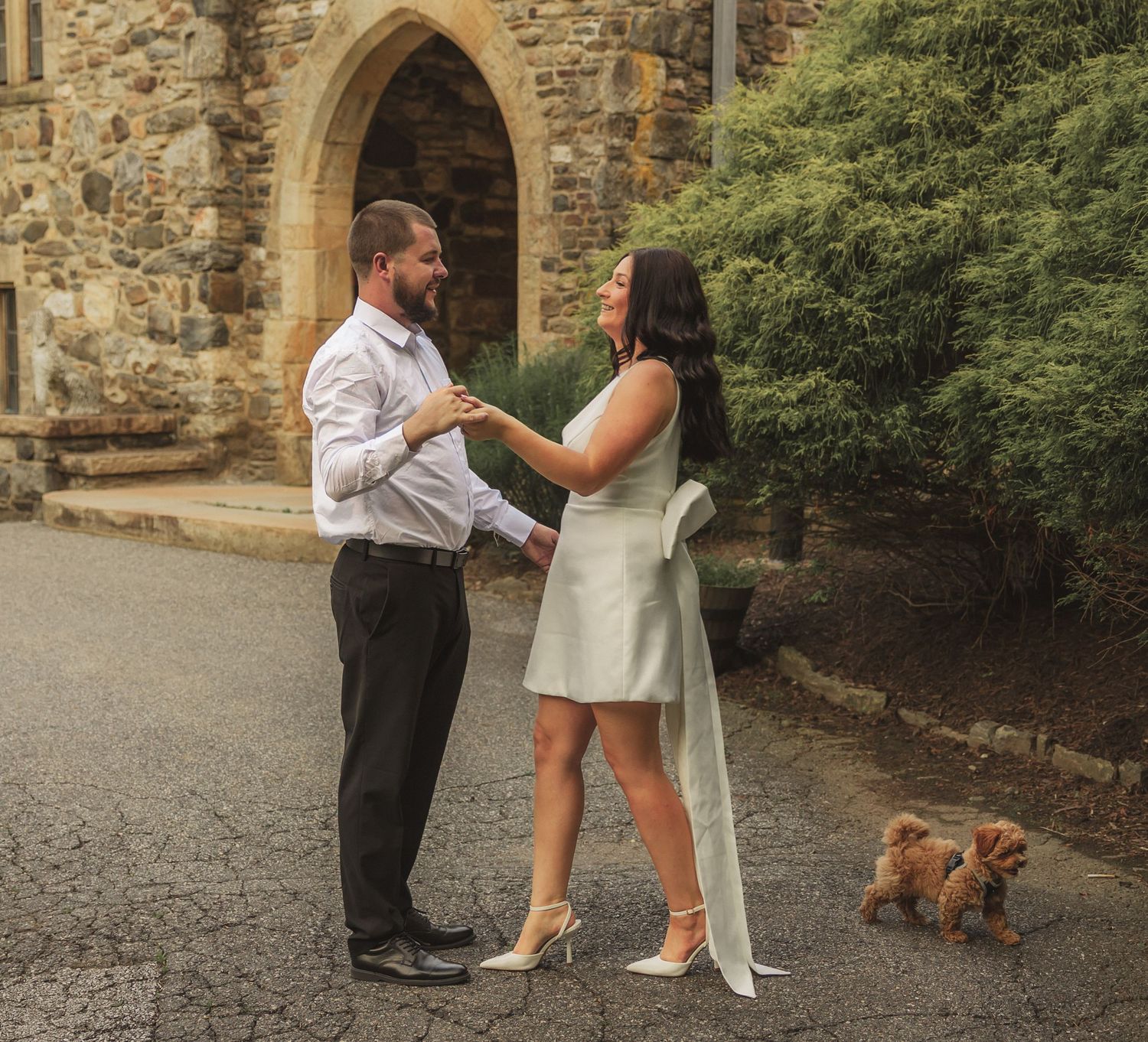 A couple shares a romantic moment outside a stone church archway while their small dog watches nearby.