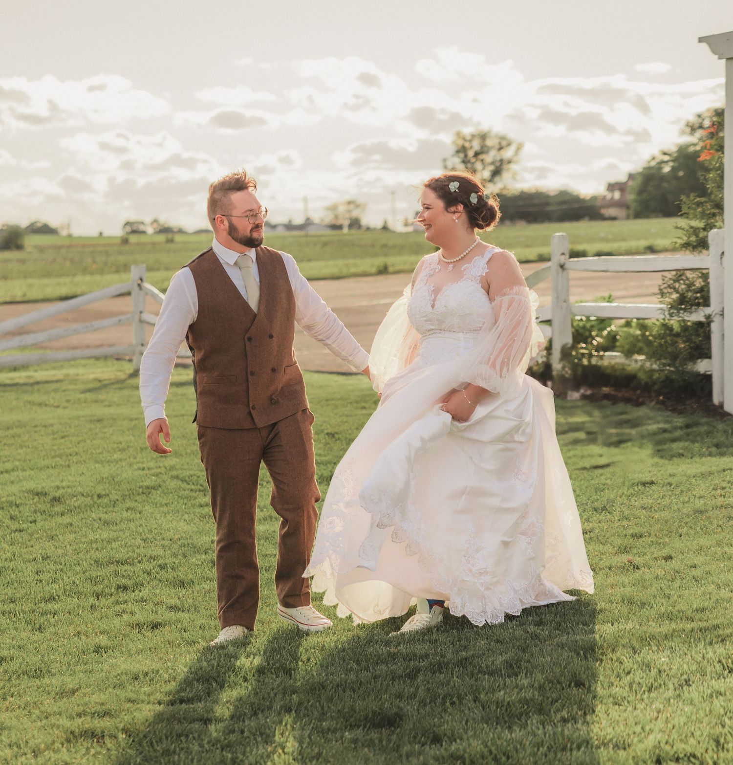 A couple in wedding attire share a joyful moment together on a grassy lawn with countryside scenery in the background.
