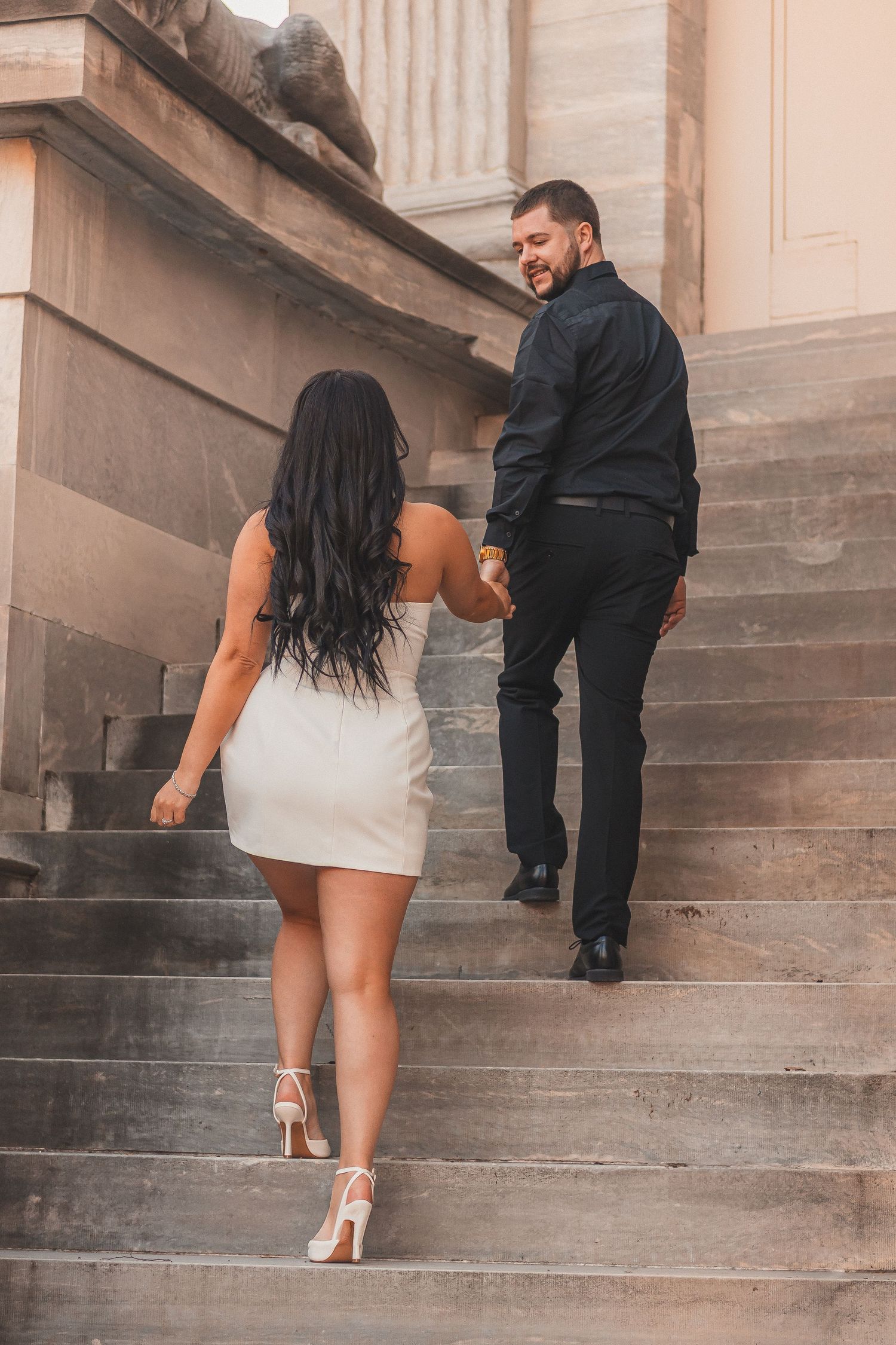 A couple ascending ornate stone stairs while holding hands and looking back at each other.