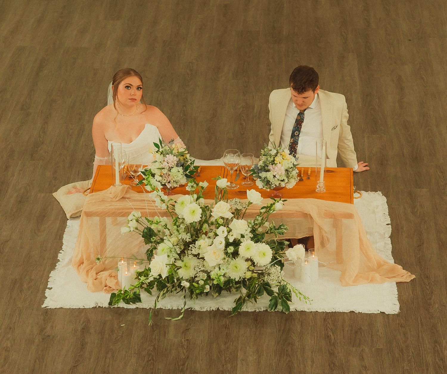 A wedding couple sits at their reception table decorated with elegant white floral arrangements and greenery.