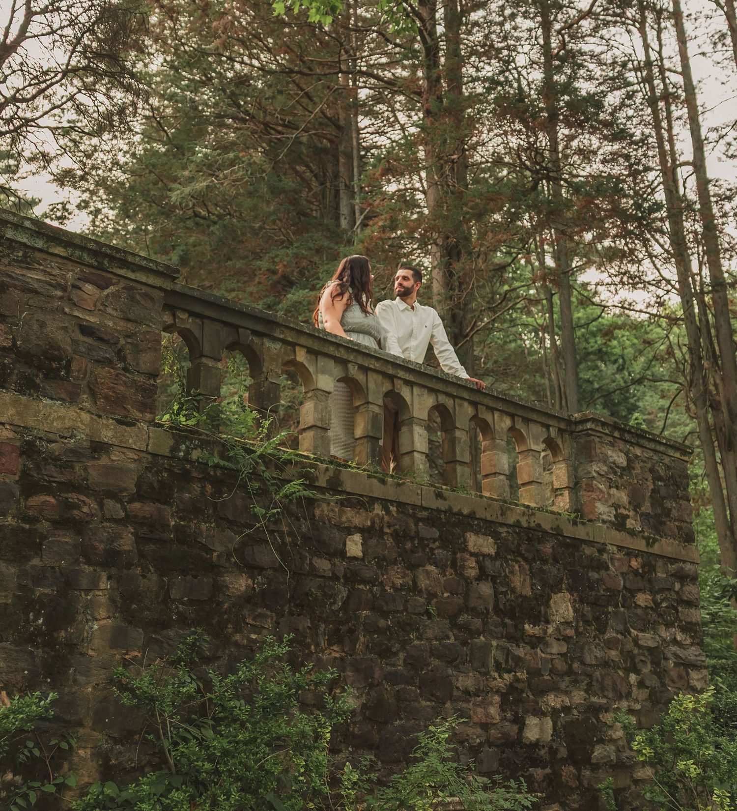 A couple stands on a rustic stone bridge overlooking a wooded area at sunset, creating a romantic setting.