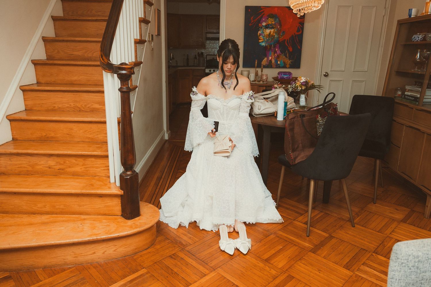 A bride in a flowing white dress and sneakers sits on wooden stairs inside a home.