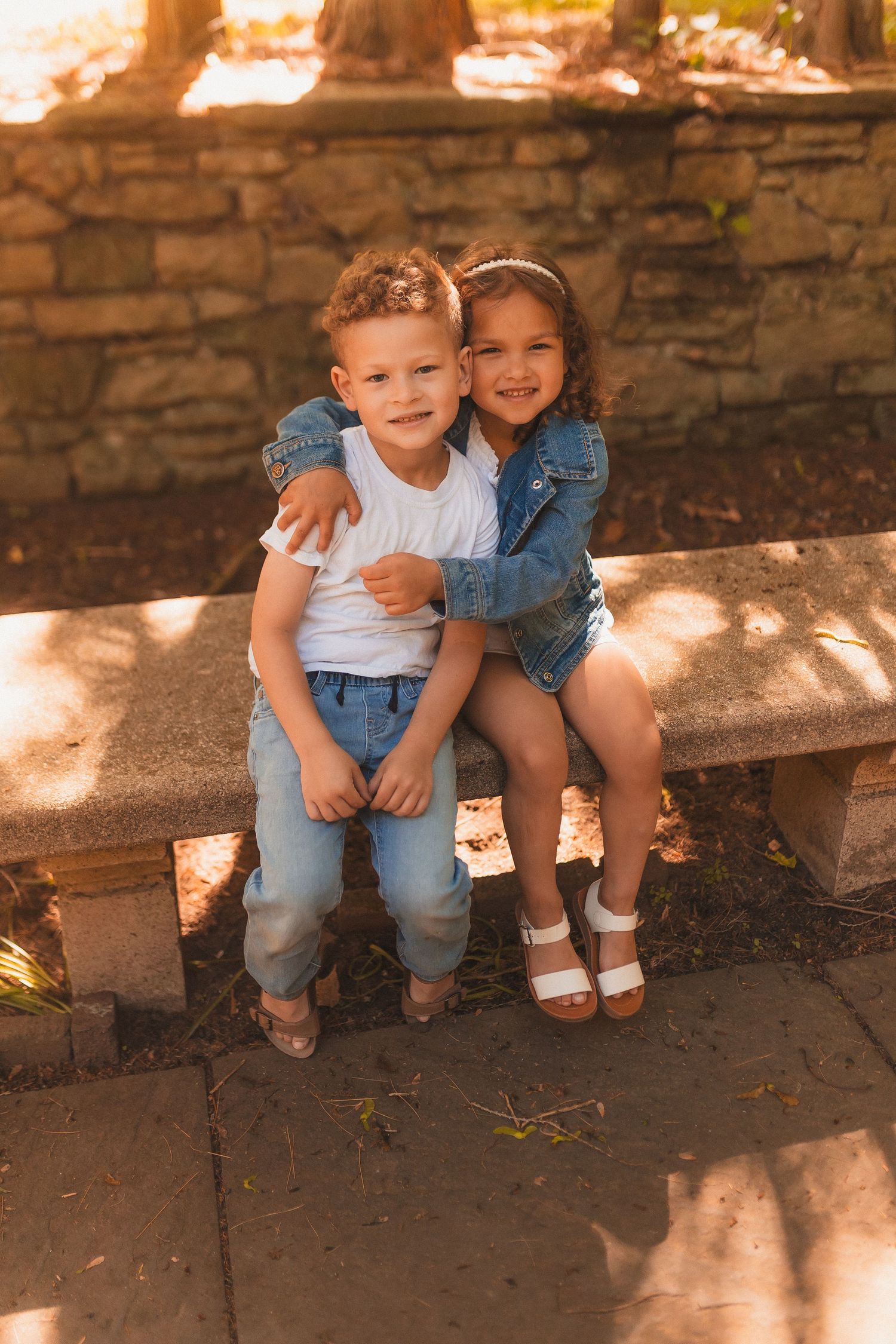 Two young children sitting together on a stone bench in dappled sunlight wearing denim and casual attire.