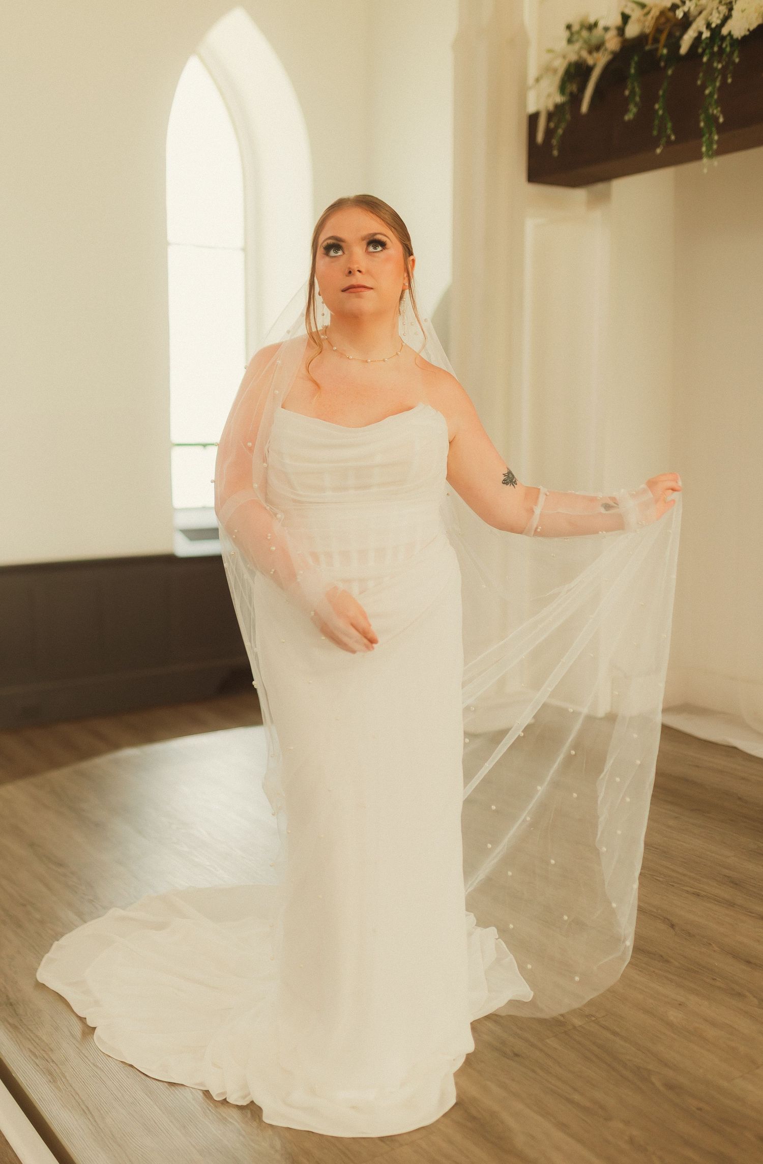 A bride in an elegant white strapless gown with flowing veil poses in front of an arched church window.