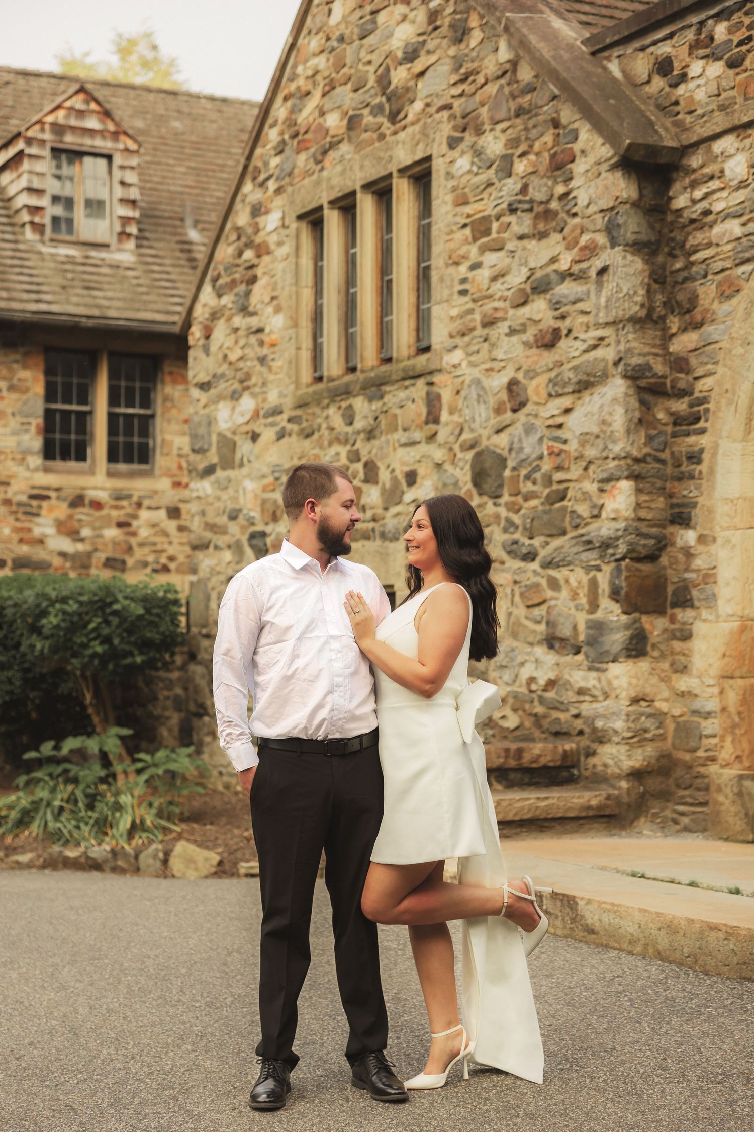 A couple in formal attire poses romantically in front of a rustic stone church building.