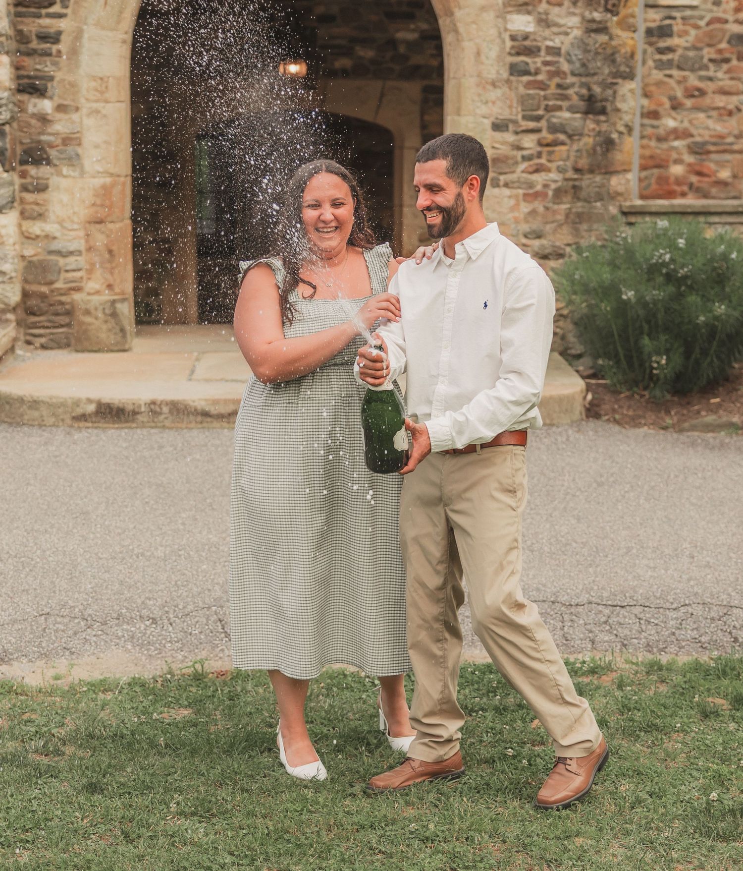 Couple celebrating with champagne spray in front of stone archway on a sunny outdoor occasion.