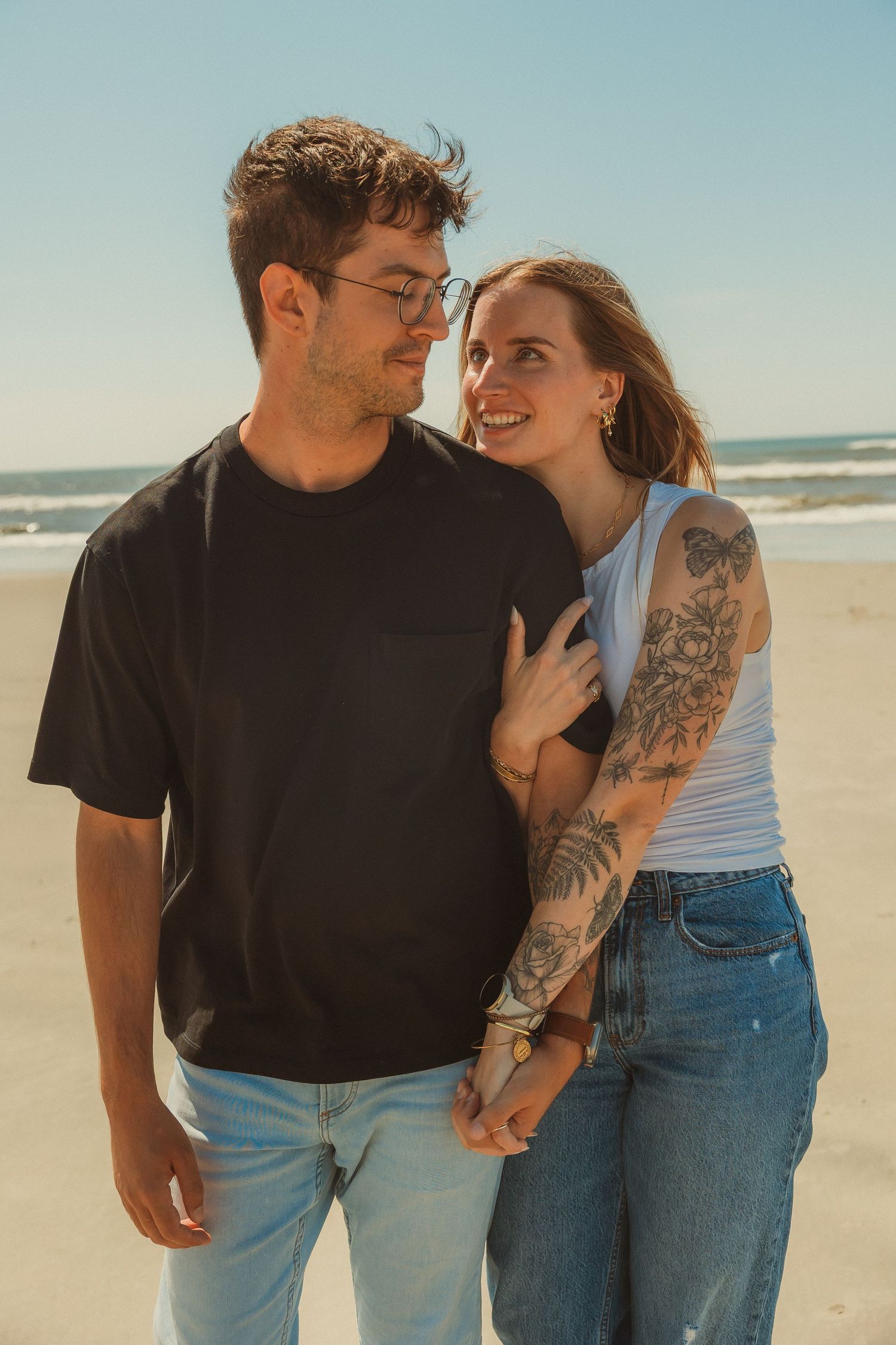A romantic couple stands together on a sunny beach, the woman wearing a white tank top with visible tattoos.