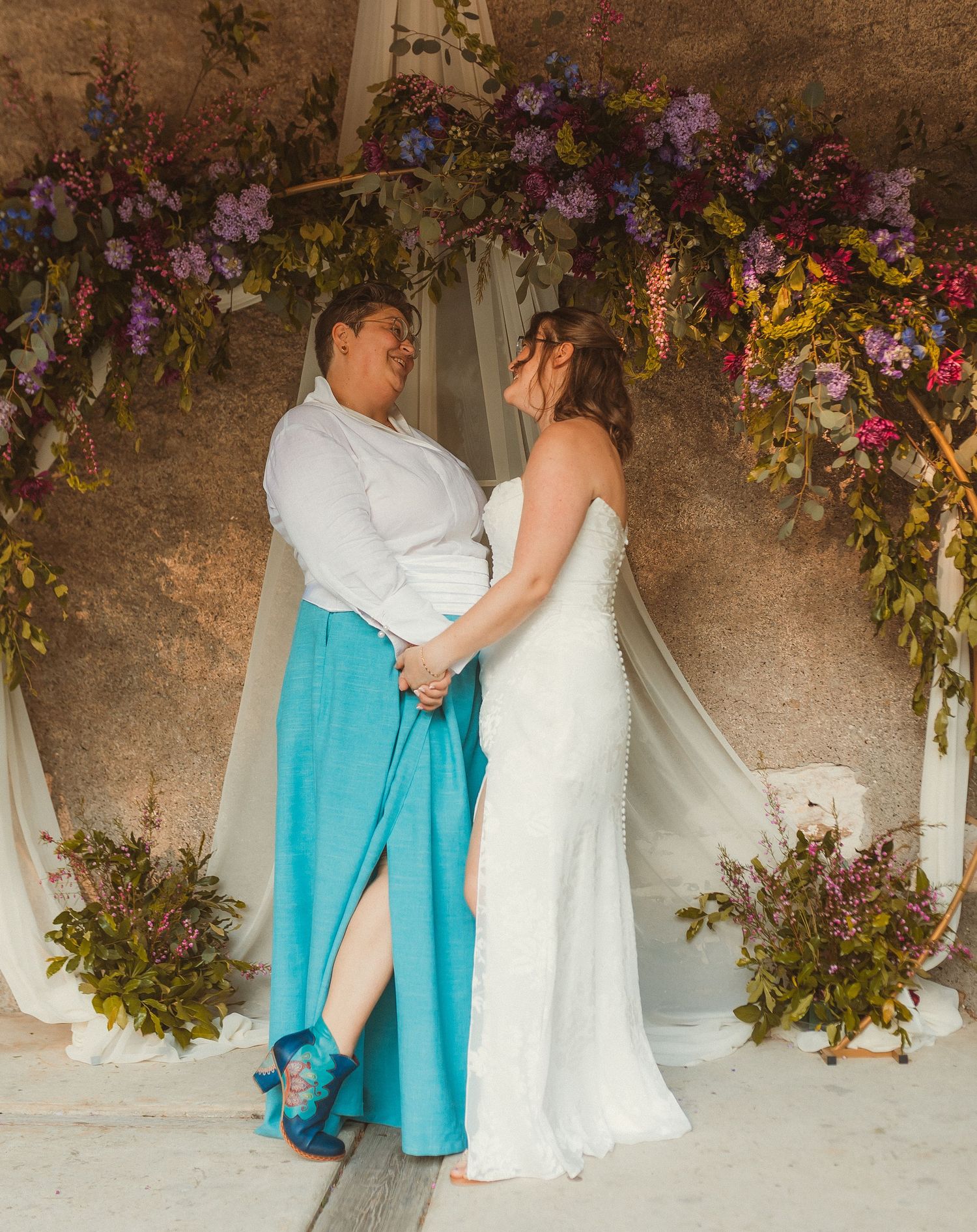 A joyful wedding couple sharing a dance under a floral arch with purple blooms.