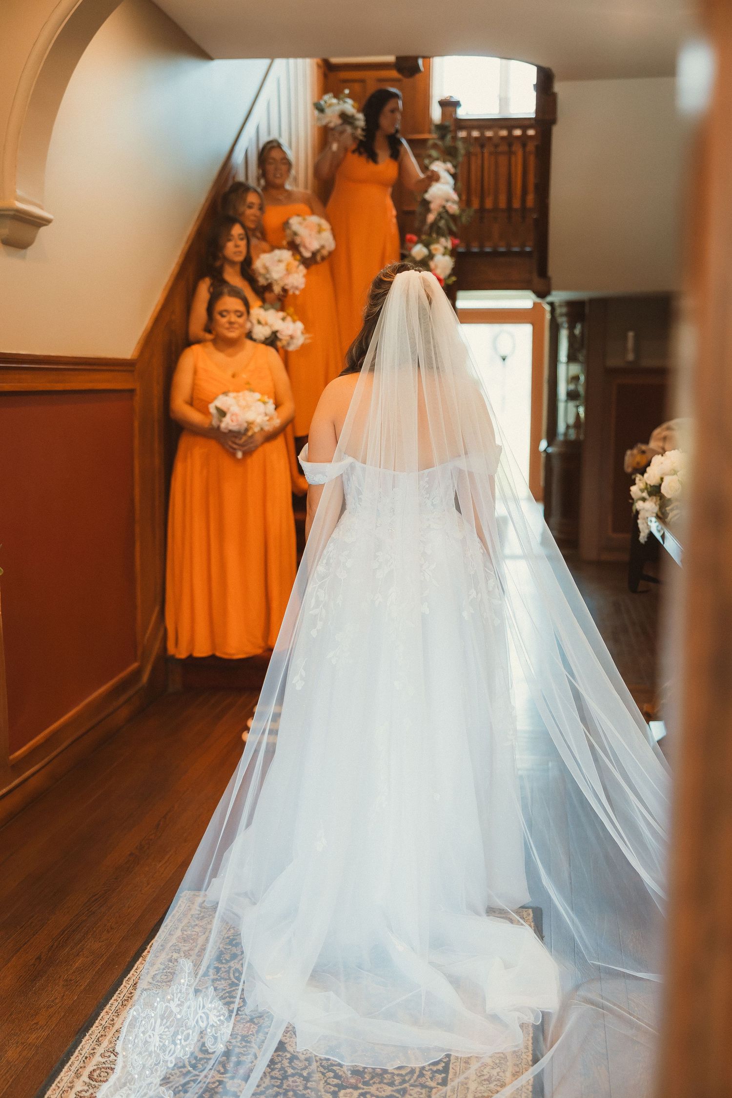 Bridal party in vibrant orange dresses lined up on wooden staircase with flowing white wedding veil.