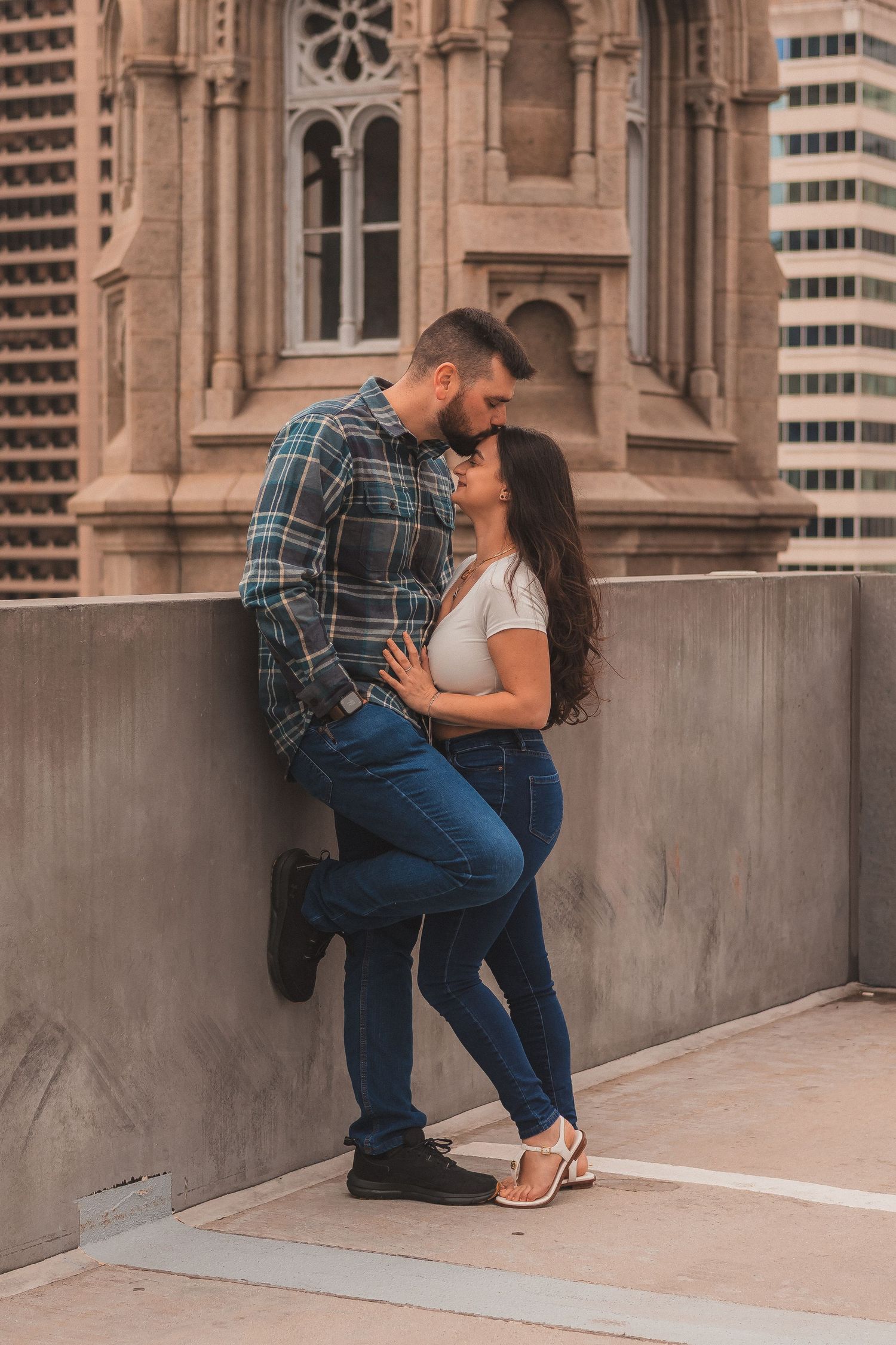 A romantic couple embraces against a historic stone building with modern skyscrapers in the background.