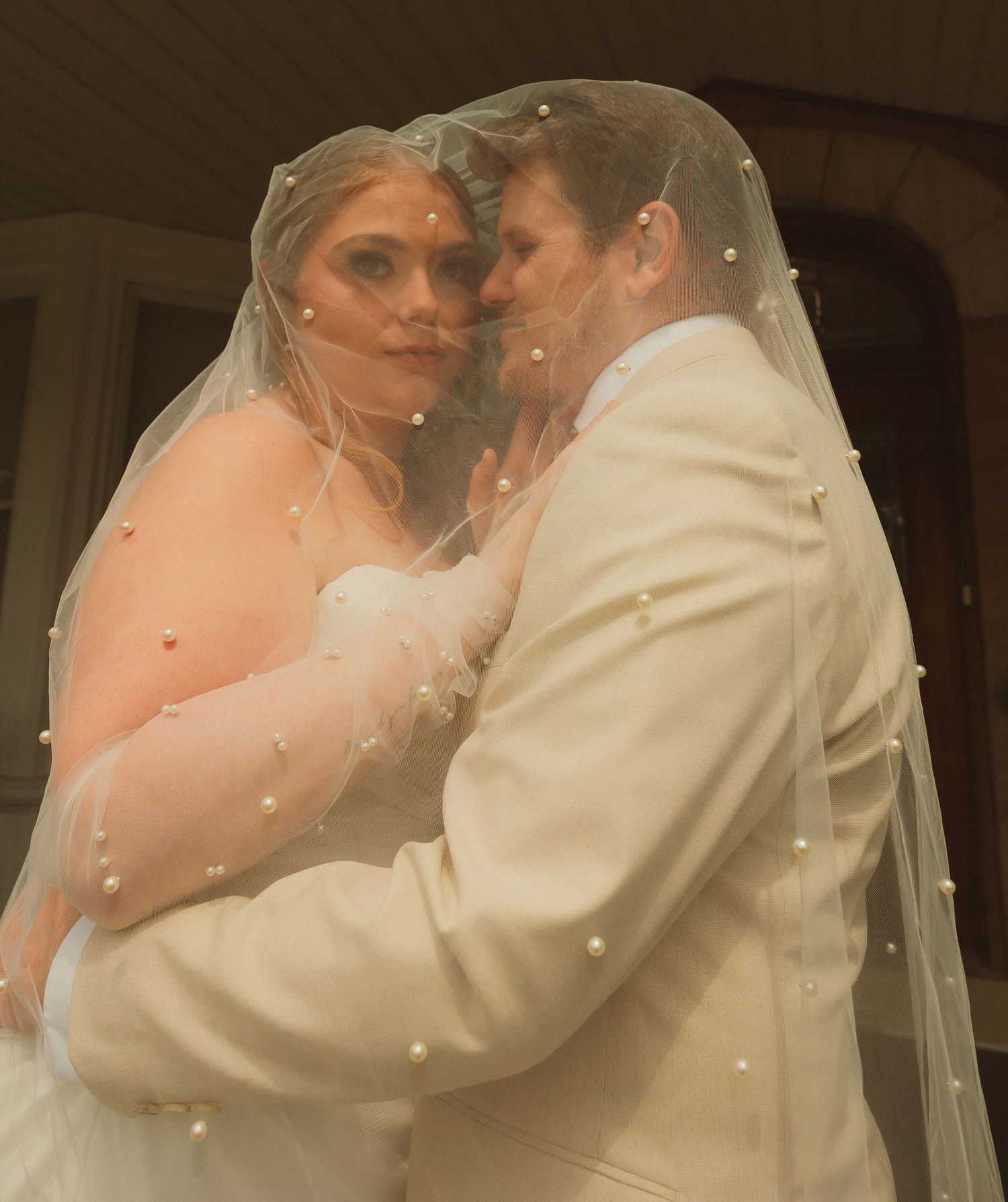 An intimate wedding portrait capturing the couple sharing a romantic moment beneath a sheer veil with pearl accents.