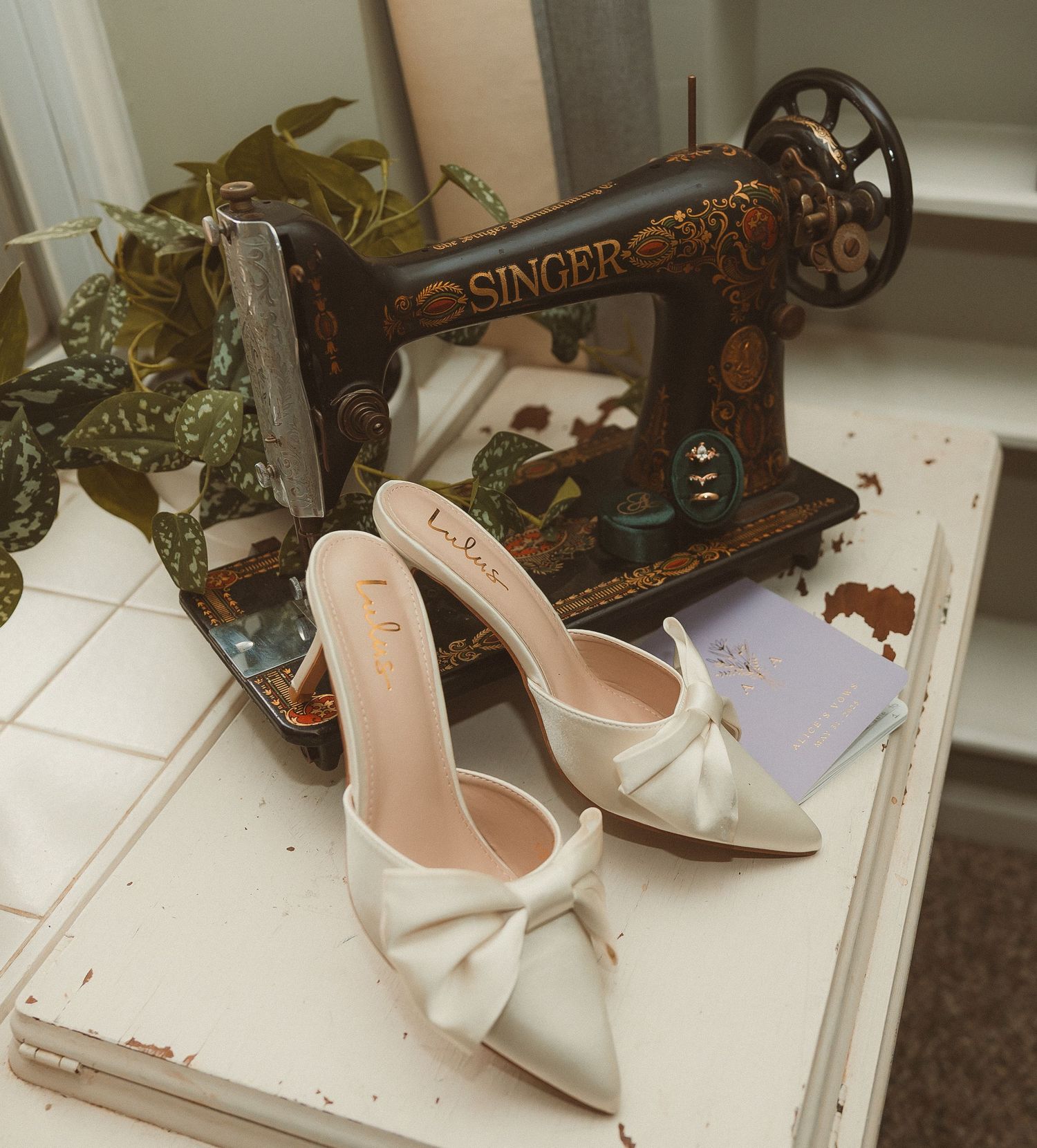 Elegant white bow heels displayed next to vintage rustic Singer sewing machine and decorative greenery.