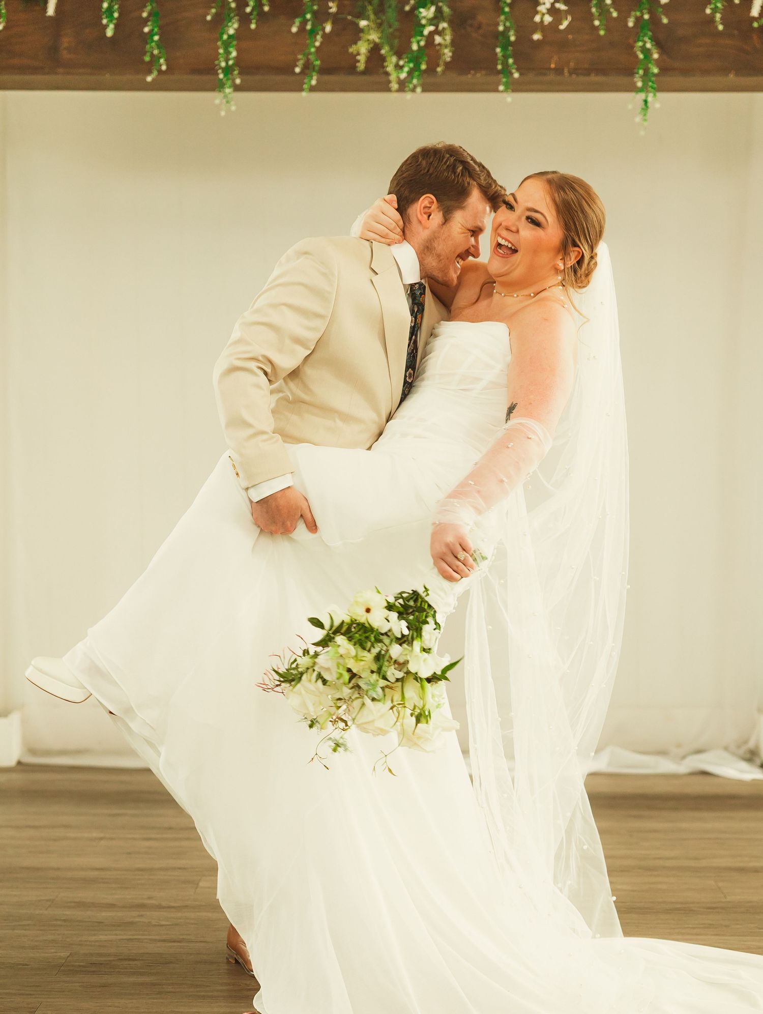 Joyful couple shares an intimate moment while dancing in a wedding dress and beige suit beneath hanging greenery.