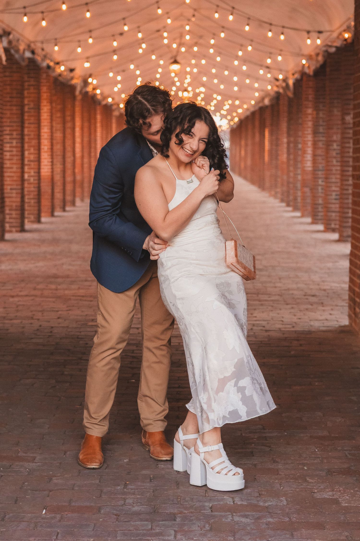 A couple shares an intimate moment in a brick corridor lit with string lights above.