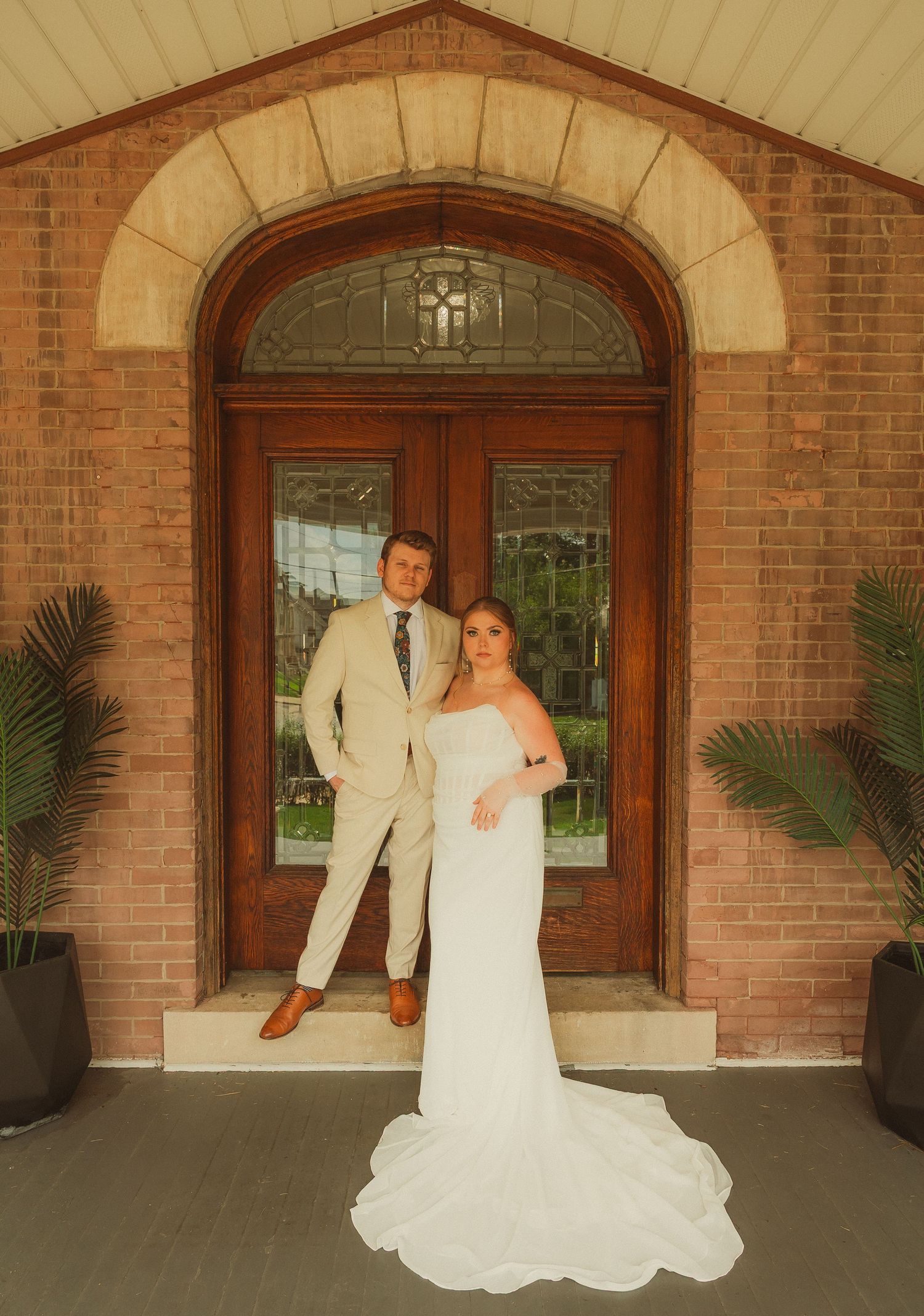 A couple stands in front of a grand arched brick doorway on their wedding day, the groom in a light suit and bride in white.