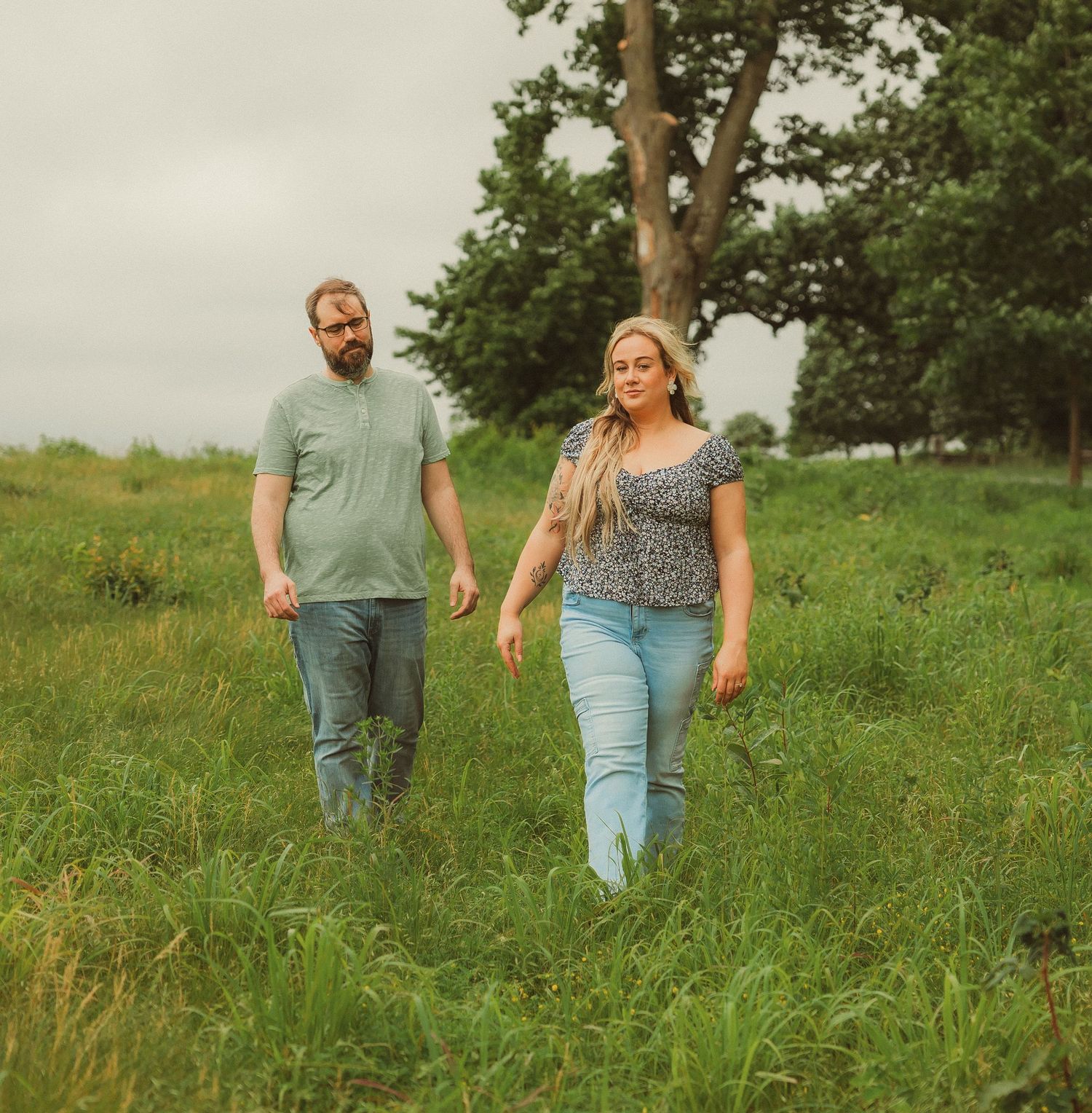 Two people walk through a lush green field on a cloudy summer day while holding hands.