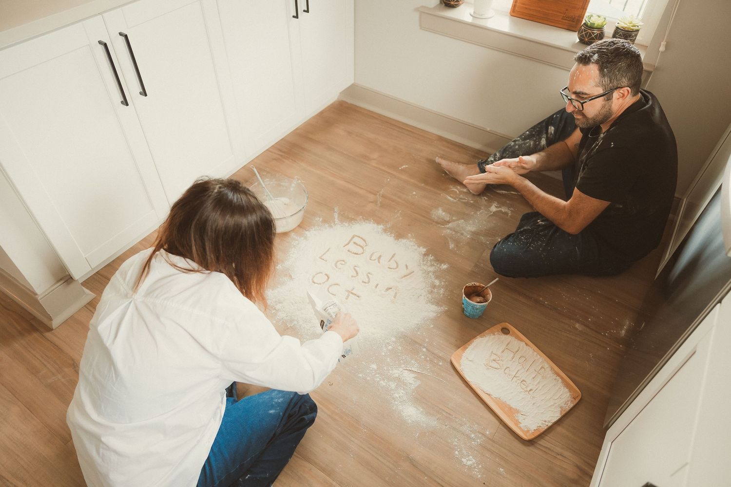 Two people sit on wooden floor playing with flour or dough in a kitchen while white powder covers the ground.