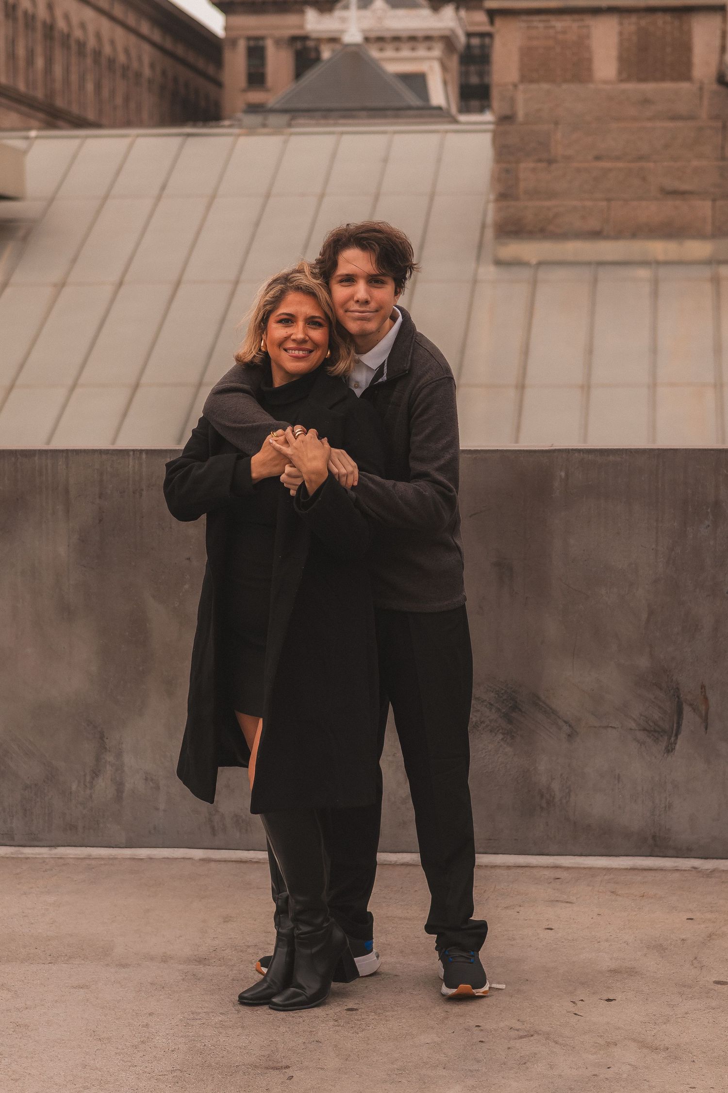 Couple in dark winter coats embracing against historic building backdrop.