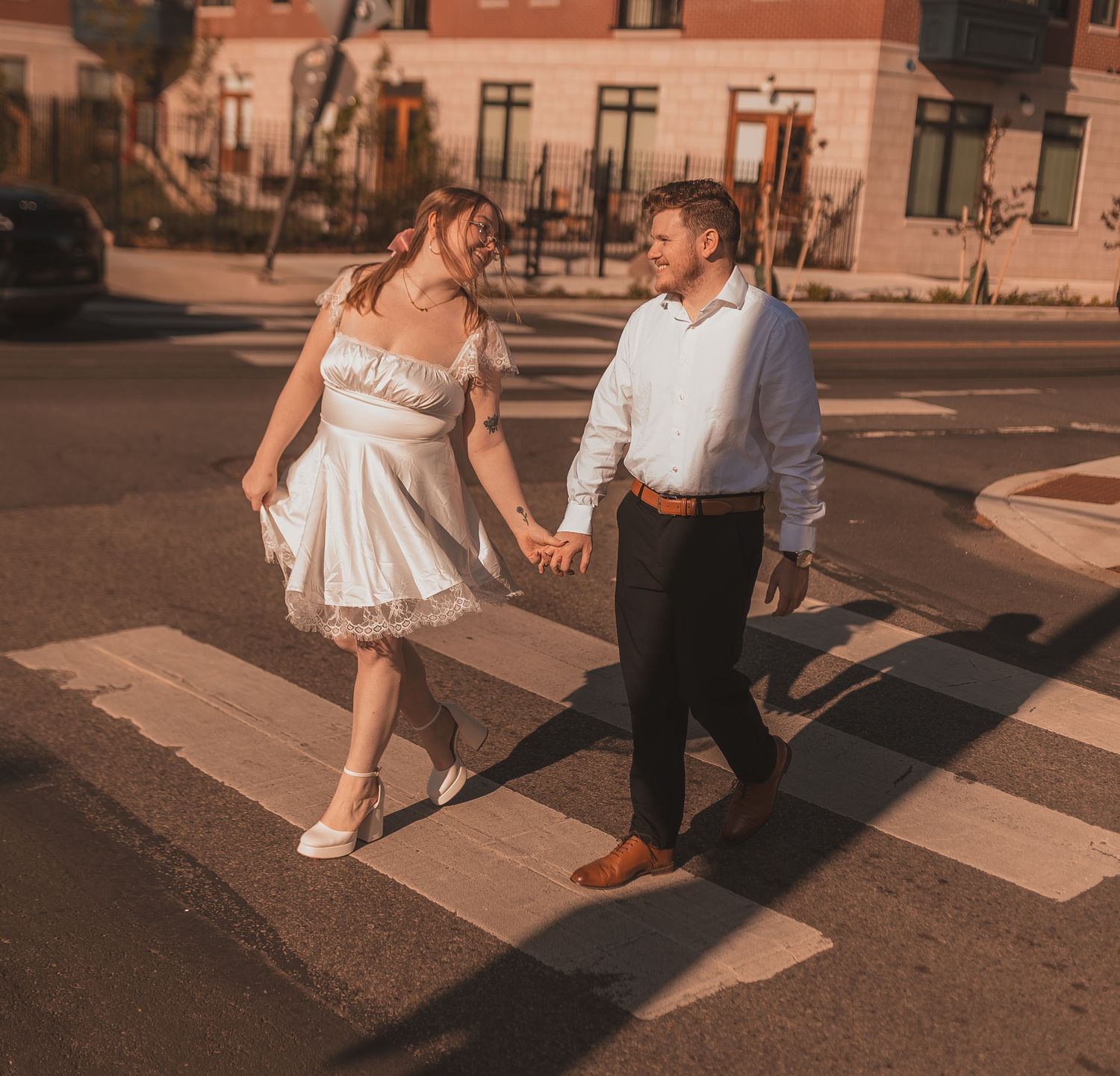 A couple in formal wear holds hands while crossing a city street at sunset.