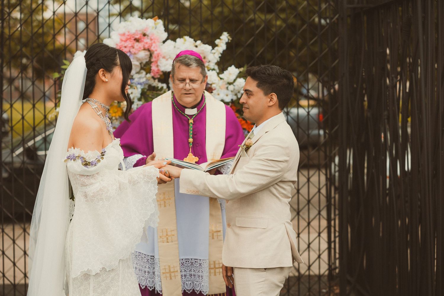 A beautiful outdoor wedding ceremony takes place with a priest in pink robes and a wooden cross under natural lighting.