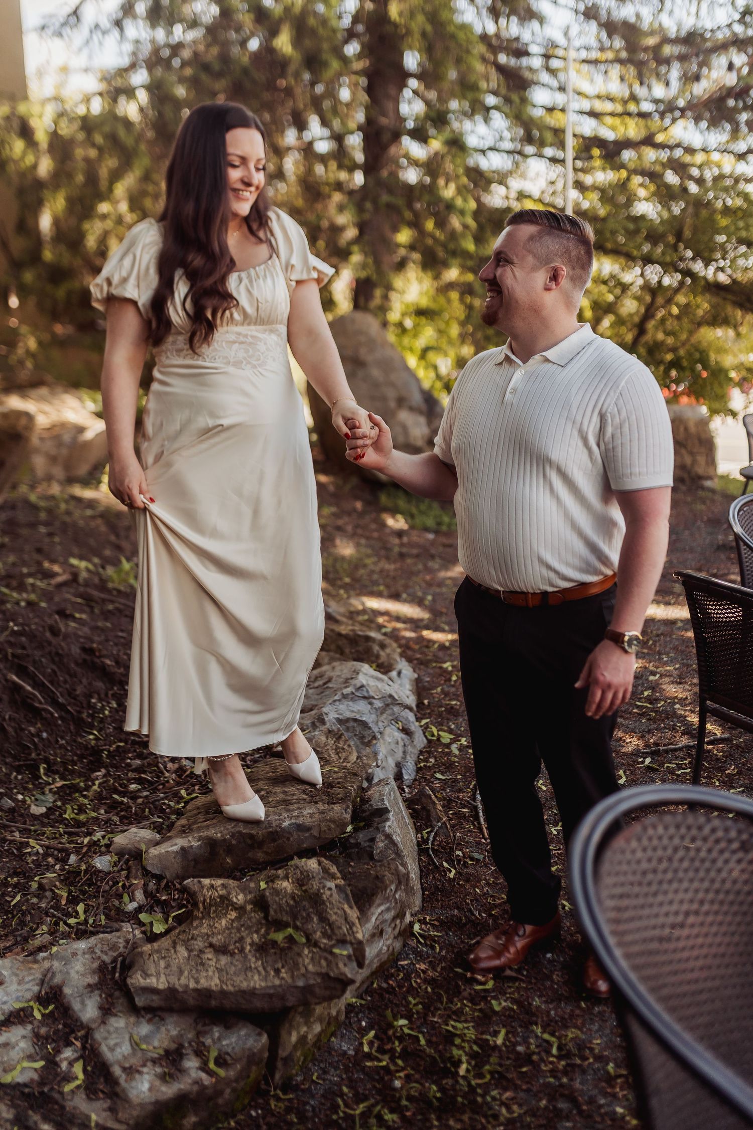A couple in casual attire share a romantic moment on rustic stone steps surrounded by evergreen trees in soft lighting.
