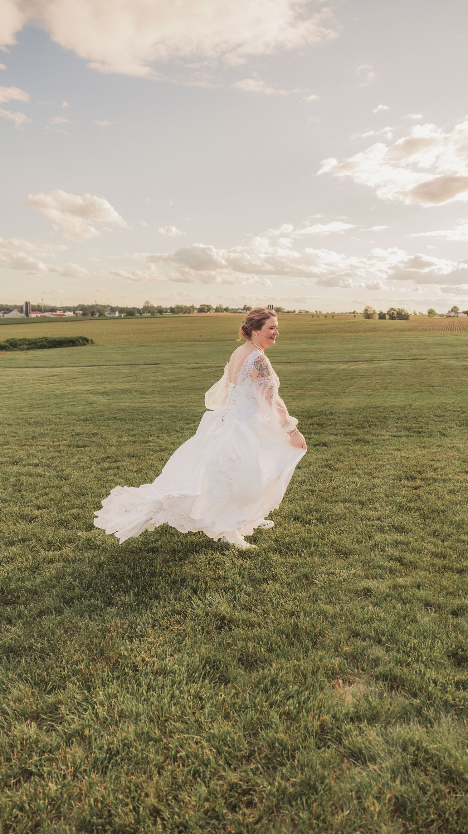 A flowing white wedding gown twirls gracefully in an open green field under a soft sunset sky.