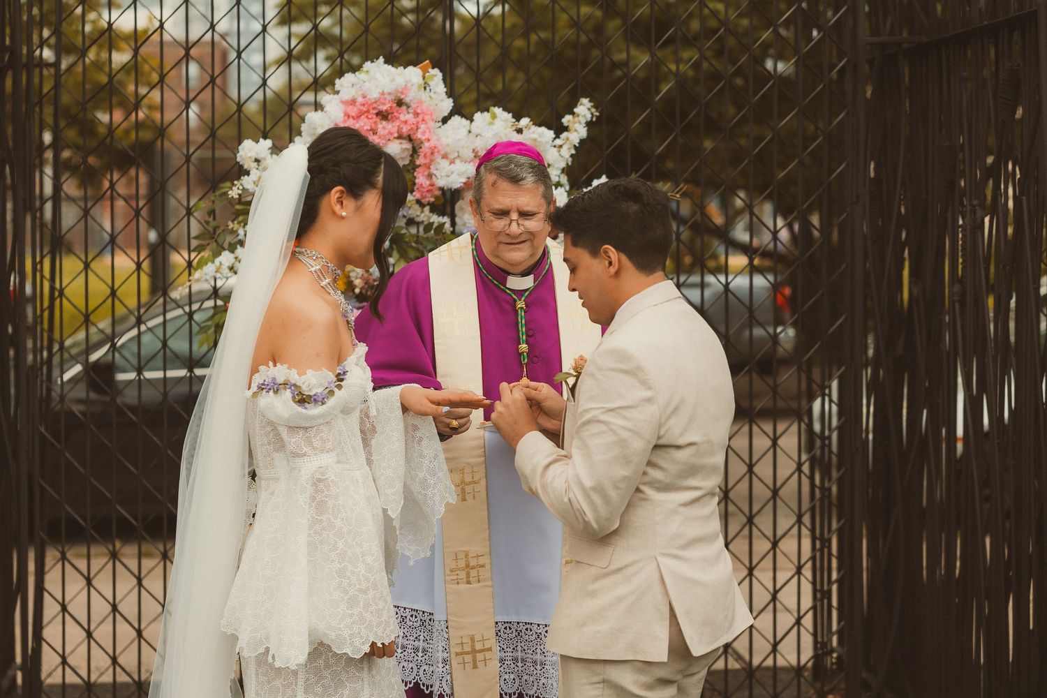 Wedding ceremony with couple and officiant in purple robe exchanging rings in front of iron gate during sunset.