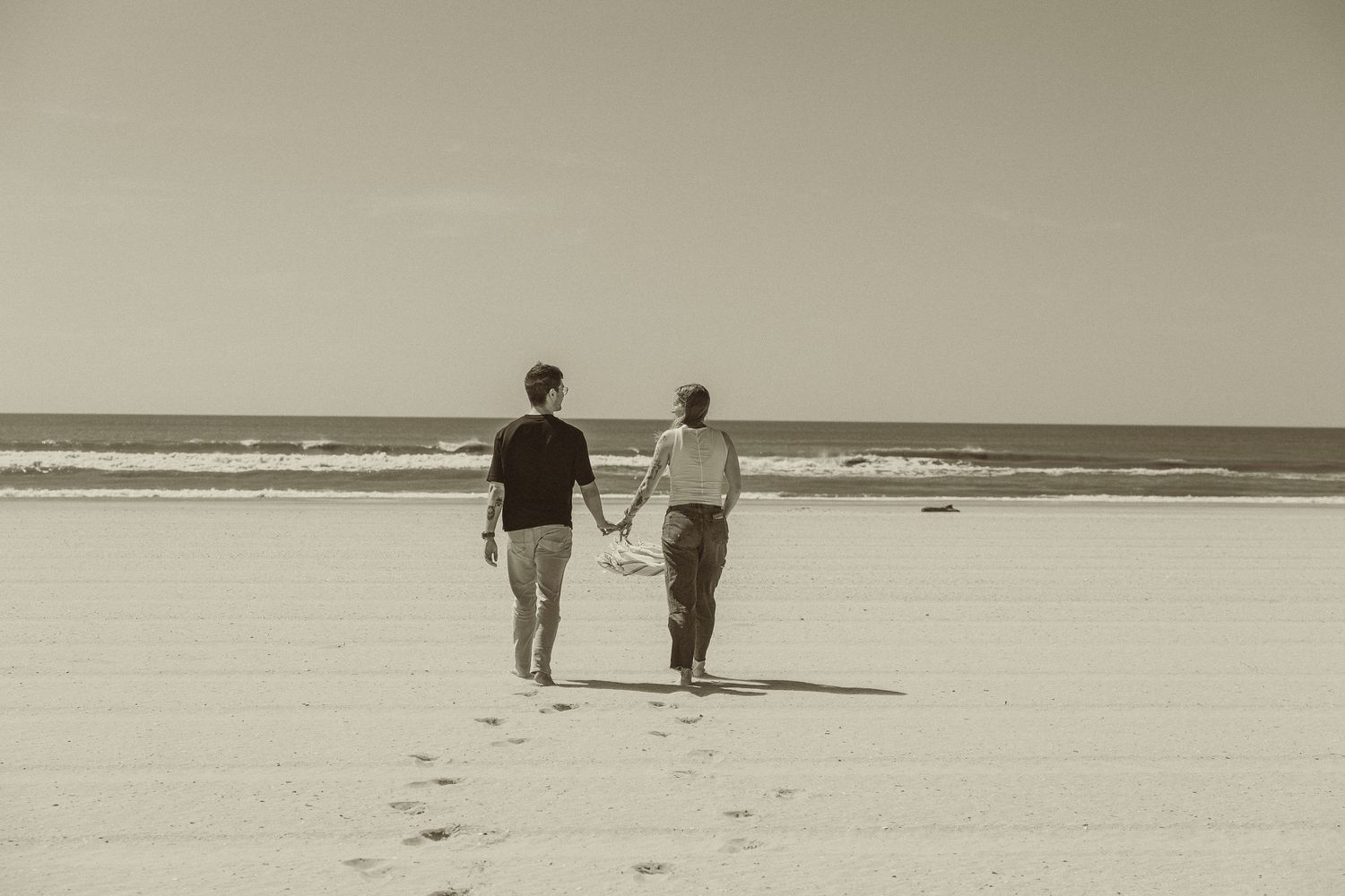 Two people walk along the sandy beach shore with ocean waves and horizon visible in a vintage sepia photograph.