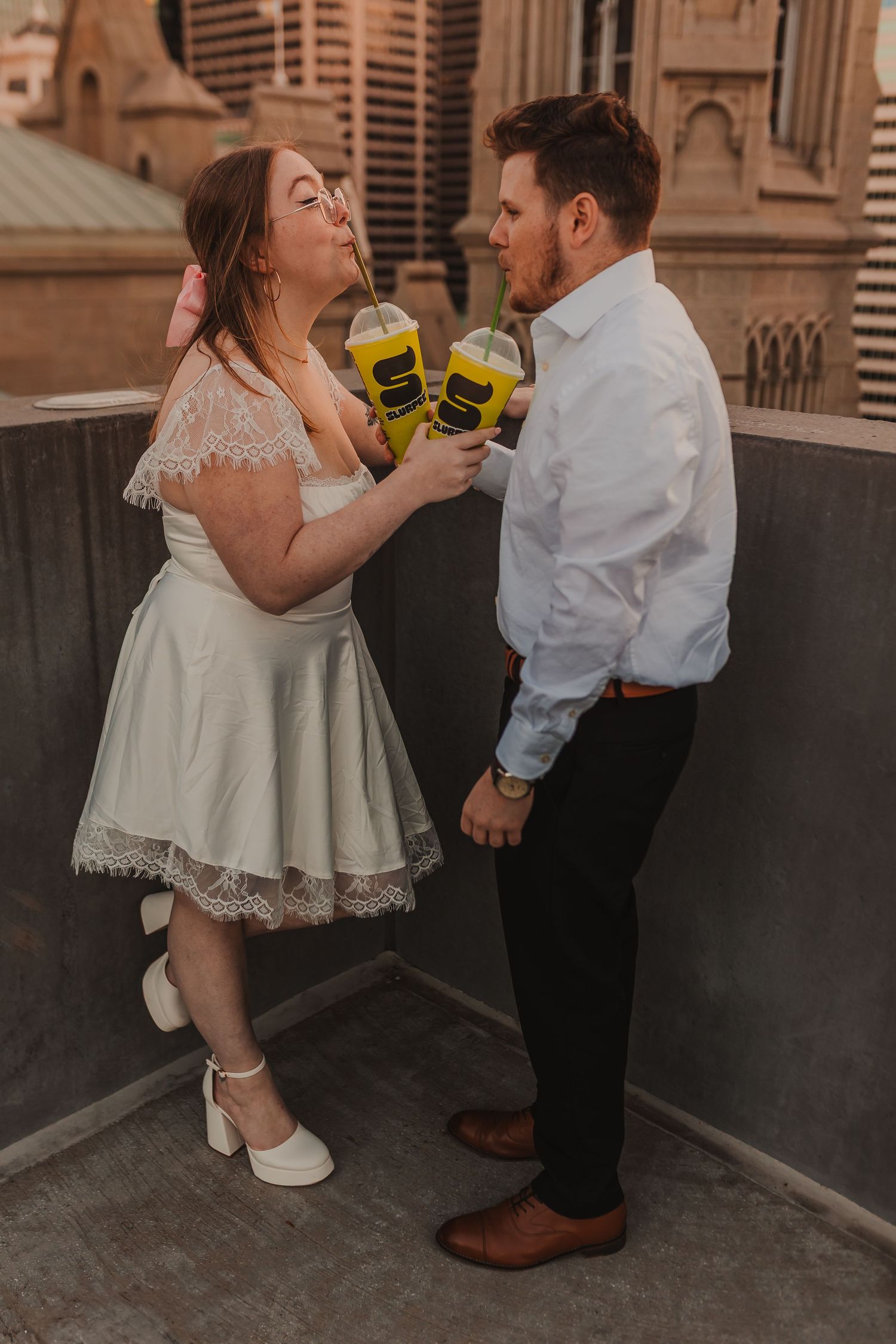 Couple playfully toasts with yellow coffee mugs on city rooftop at sunset.