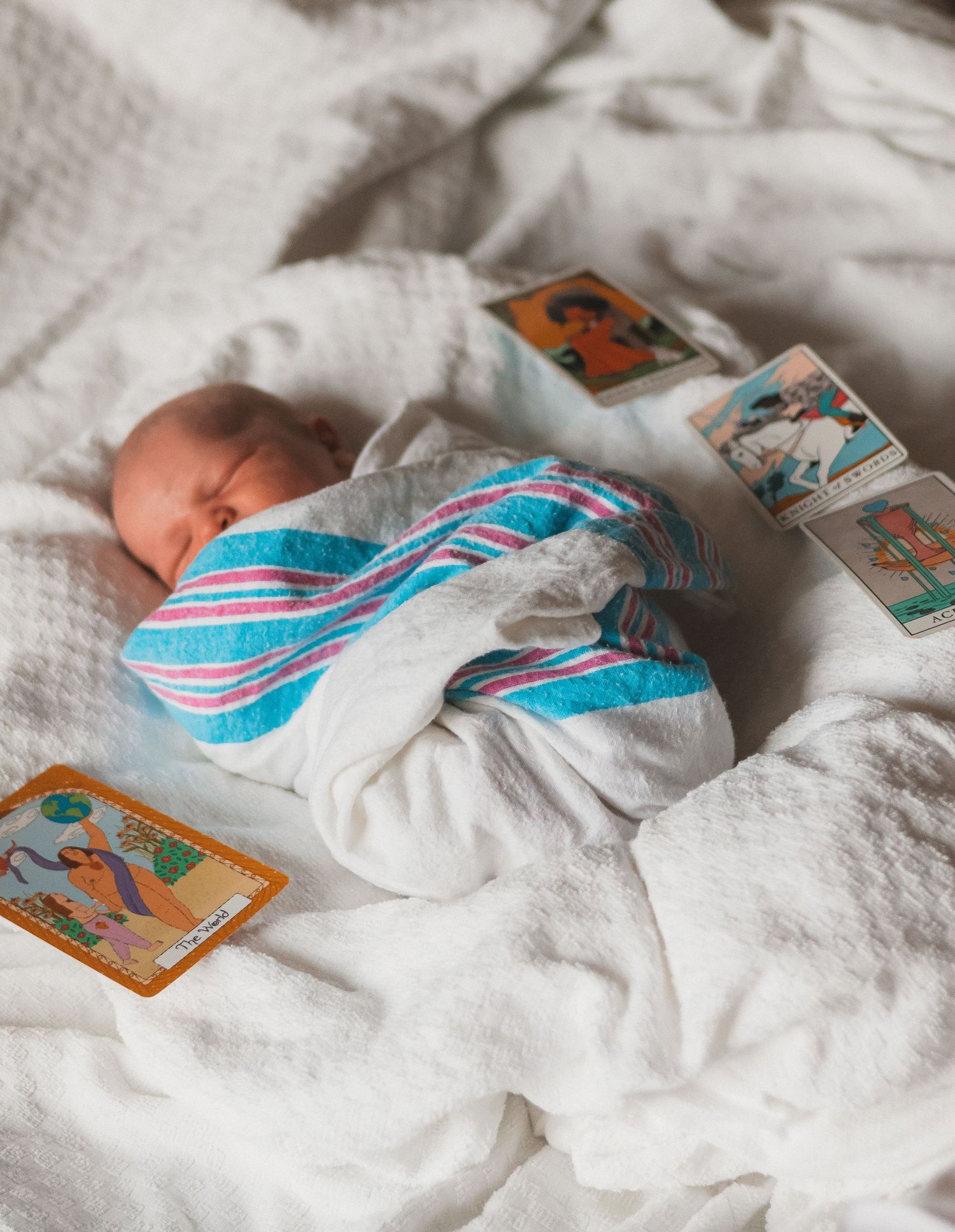 A newborn baby swaddled in a turquoise and white blanket laying on white bedding with playing cards nearby.