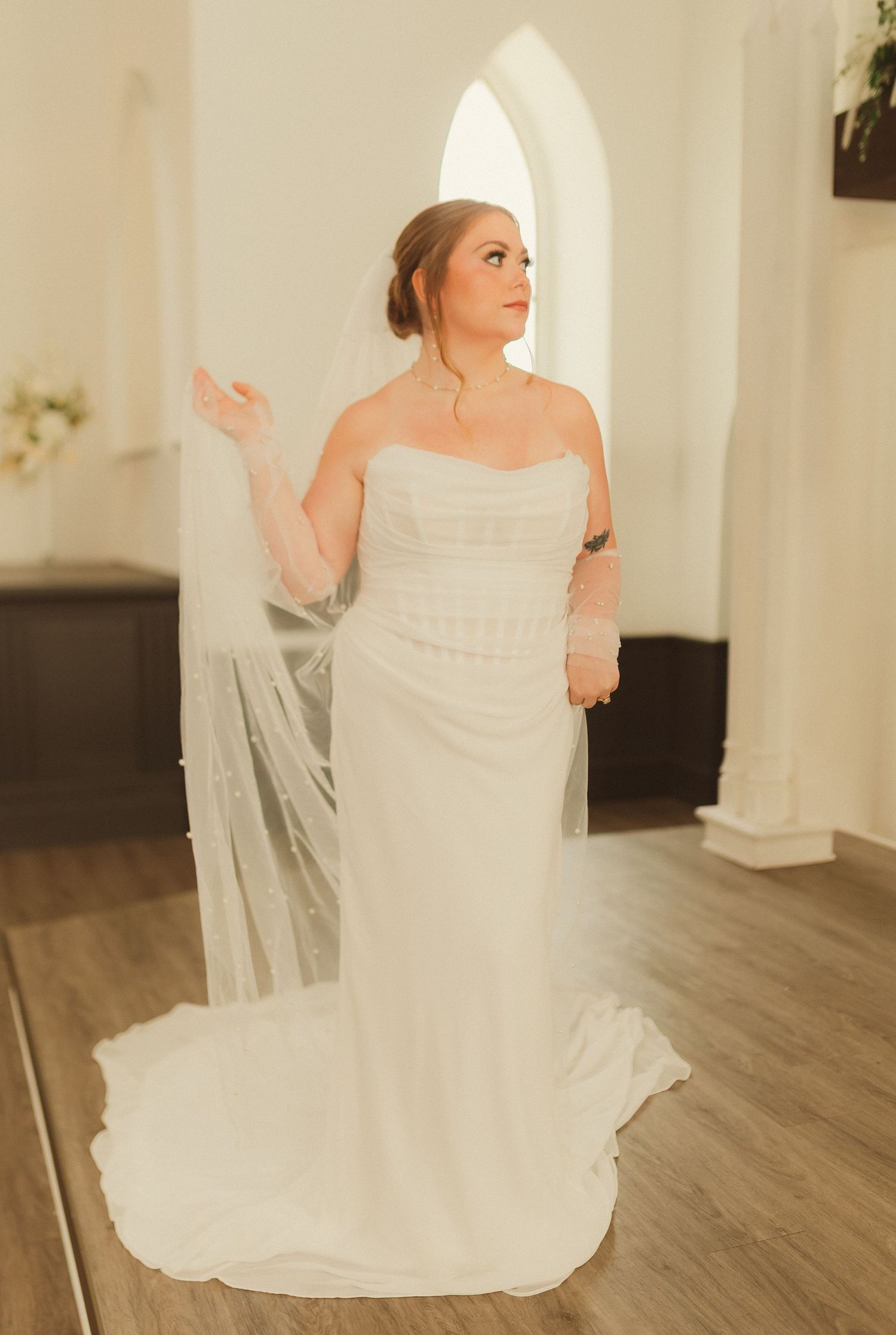 A bride in a flowing white strapless wedding dress with train stands gracefully in a room with arched windows and hardwood floors.