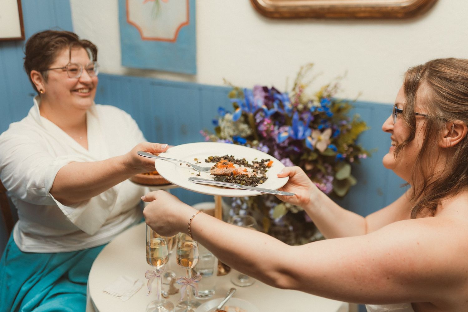 Two people sharing a plate of food at a celebration with blue flowers in the background.