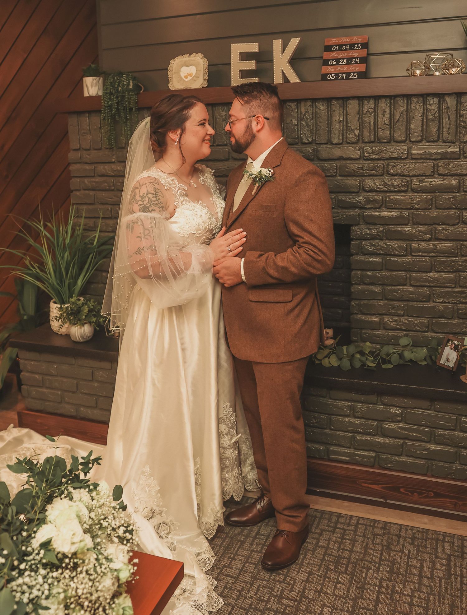 A couple in wedding attire share a romantic moment outside against a rustic brick wall with decorative lighting.